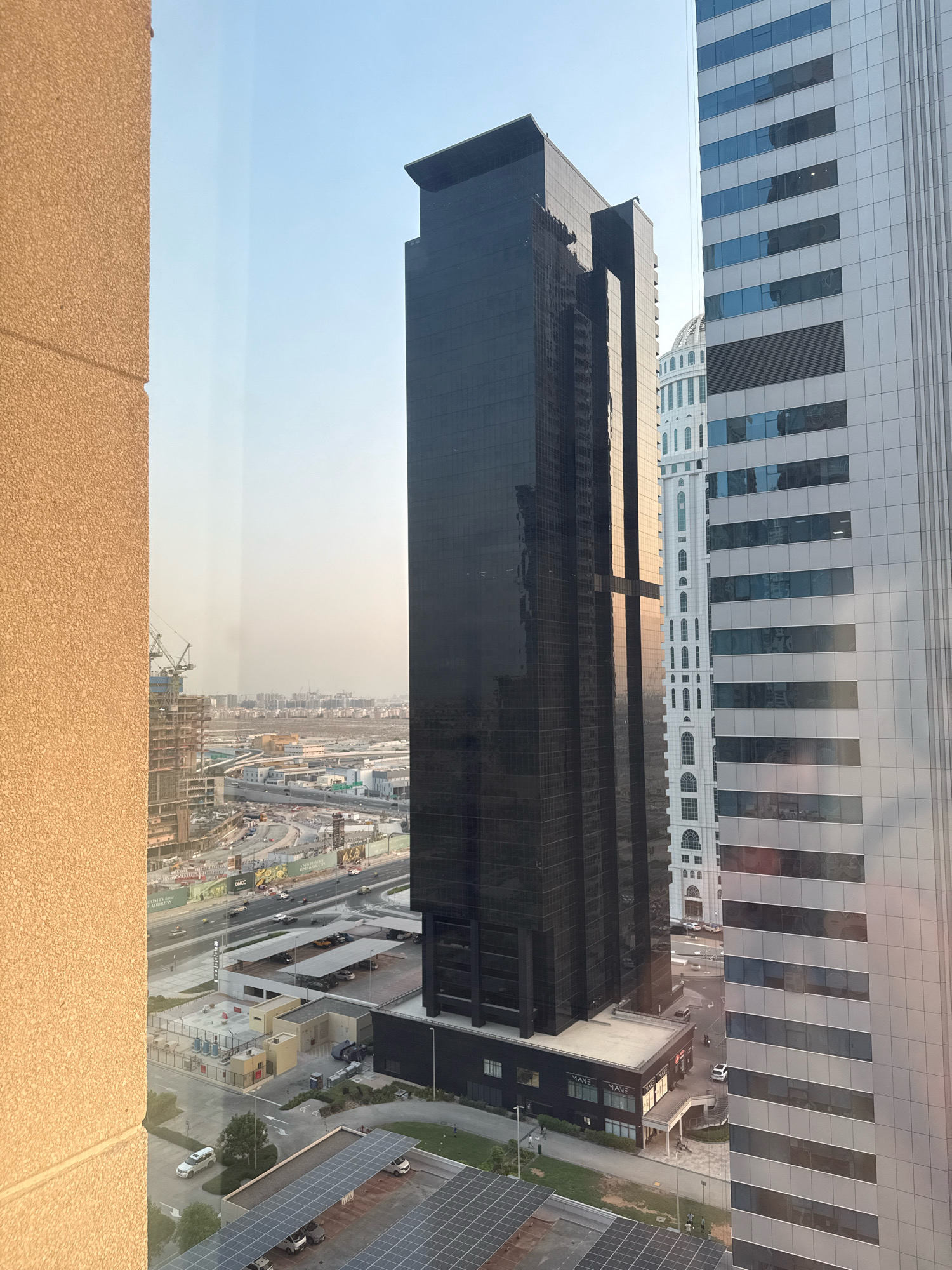 A high-angle, outdoor, full shot captures a cityscape in Dubai, United Arab Emirates, dominated by modern skyscrapers. In the foreground, on the left, a textured, beige building partially frames the view. To its right, a sleek, black skyscraper stands tall and imposing, its glass facade reflecting the sky. Adjacent to it, a white building with arched windows adds architectural contrast, and further to the right, a predominantly gray and white skyscraper with horizontal window bands extends upwards.
Below the towering structures, a bustling urban environment unfolds. A multi-lane road snakes through the scene, with several vehicles, including cars and what appear to be taxis, navigating the traffic. Construction sites are visible, characterized by cranes and partially built structures, indicative of ongoing development. Parking lots with cars are also present, some covered by solar panels, suggesting a focus on renewable energy. A few individuals are seen walking on sidewalks and in open spaces. The sky is clear and bright, suggesting daylight conditions, possibly late afternoon given the warm light. The overall impression is one of a dynamic, rapidly evolving metropolis.