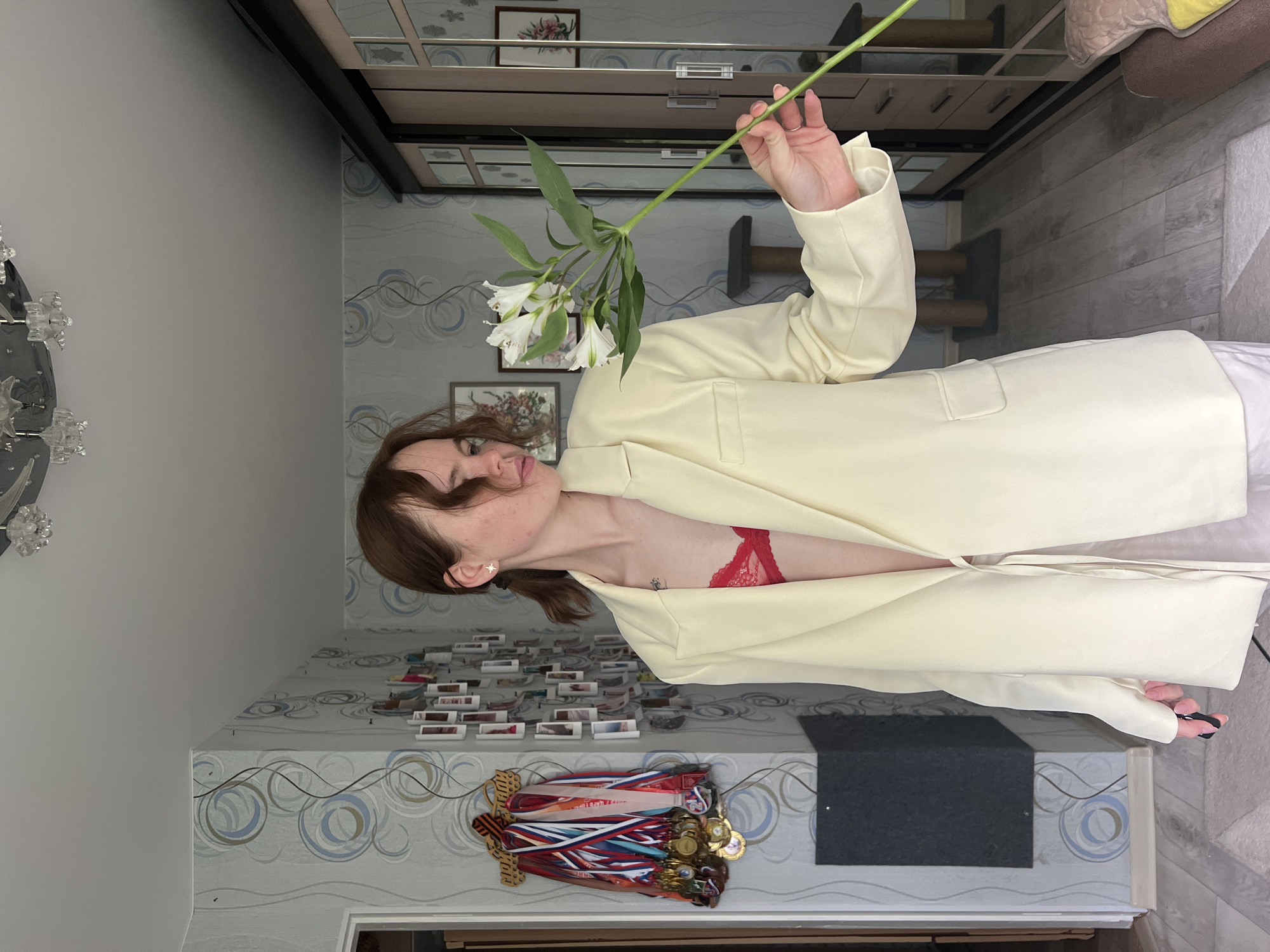A young adult female is centrally positioned within an indoor residential room in the city of Chaikovsky, Russia. She has light brown hair styled into two short ponytails and wears a light yellow, open blazer over a red lace bralette, revealing a small tattoo on her upper left chest. A star-shaped earring is visible in her right ear. She holds a stem of white alstroemeria flowers in her right hand, angled across her body, while her gaze is directed downward and to her right. Her left hand partially grasps a small black object.
The room's walls are covered with light blue-grey wallpaper featuring a repeating pattern of white, blue, and brown concentric circles and wavy lines. On the left wall, a wooden rack displays numerous sports medals with multi-colored ribbons. Below the medals, a dark grey rectangular panel is mounted. The back wall exhibits a grid arrangement of multiple small framed photographs. Two larger framed artworks depicting floral arrangements are also present, one visibly on the left and another reflected in the wardrobe on the right. A large, dark-framed sliding door wardrobe with reflective panels occupies the right side of the room. A brown cat scratching post is visible on the floor in the mid-ground. The ceiling is white, from which a decorative chrome and clear glass chandelier is suspended. The floor is comprised of light wood laminate planks, partially covered by a white and grey patterned rug in the foreground.