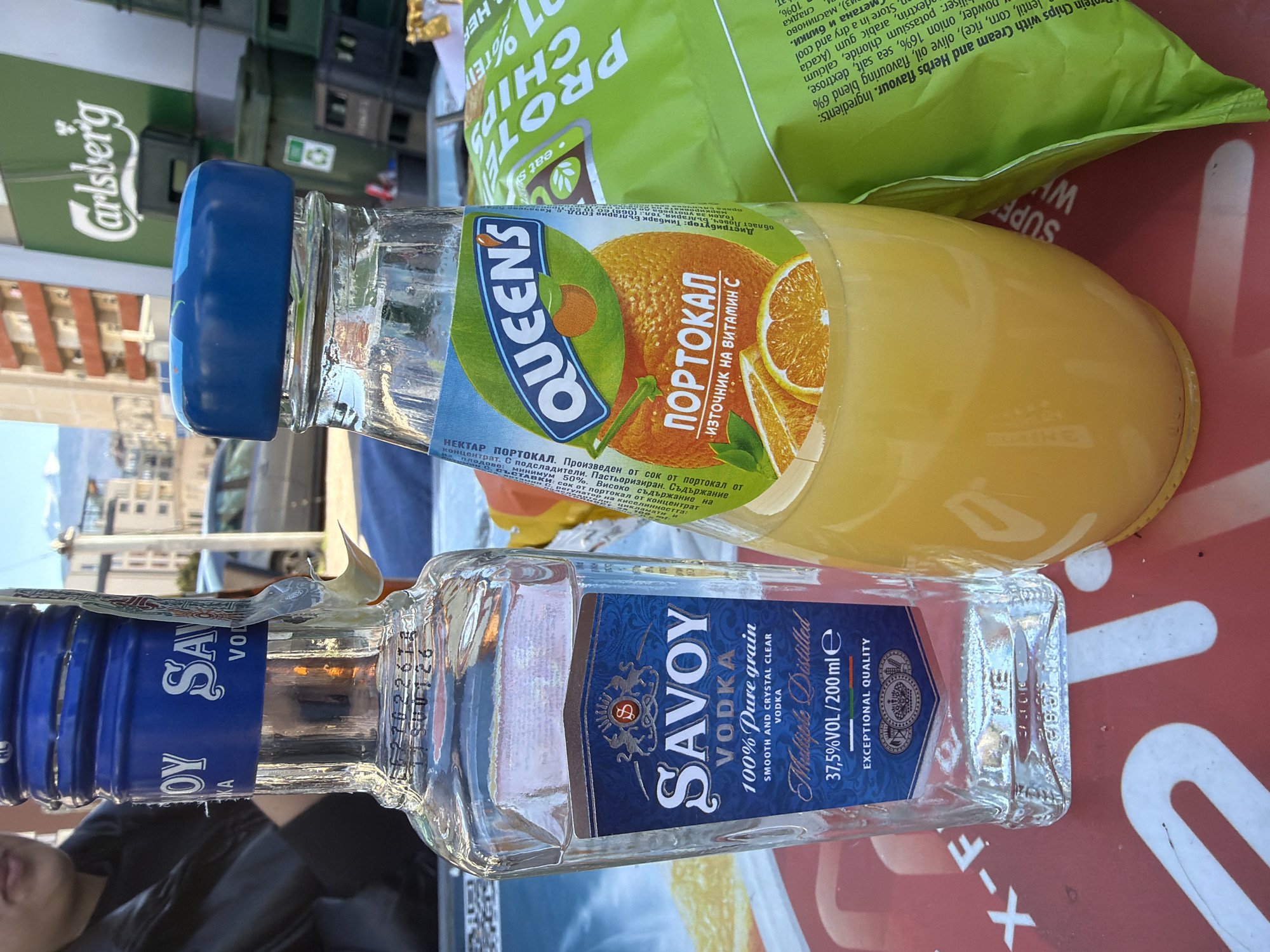 The image depicts a scene in Sofia, Bulgaria. A bottle of Savoy vodka, a bottle of Queens orange juice, and a bag of protein chips sit on a table. The vodka bottle is clear glass with a blue label and cap. The orange juice bottle is clear with a blue cap and a label featuring an orange. The protein chips bag is green. A Carlsberg sign is partially visible in the background.