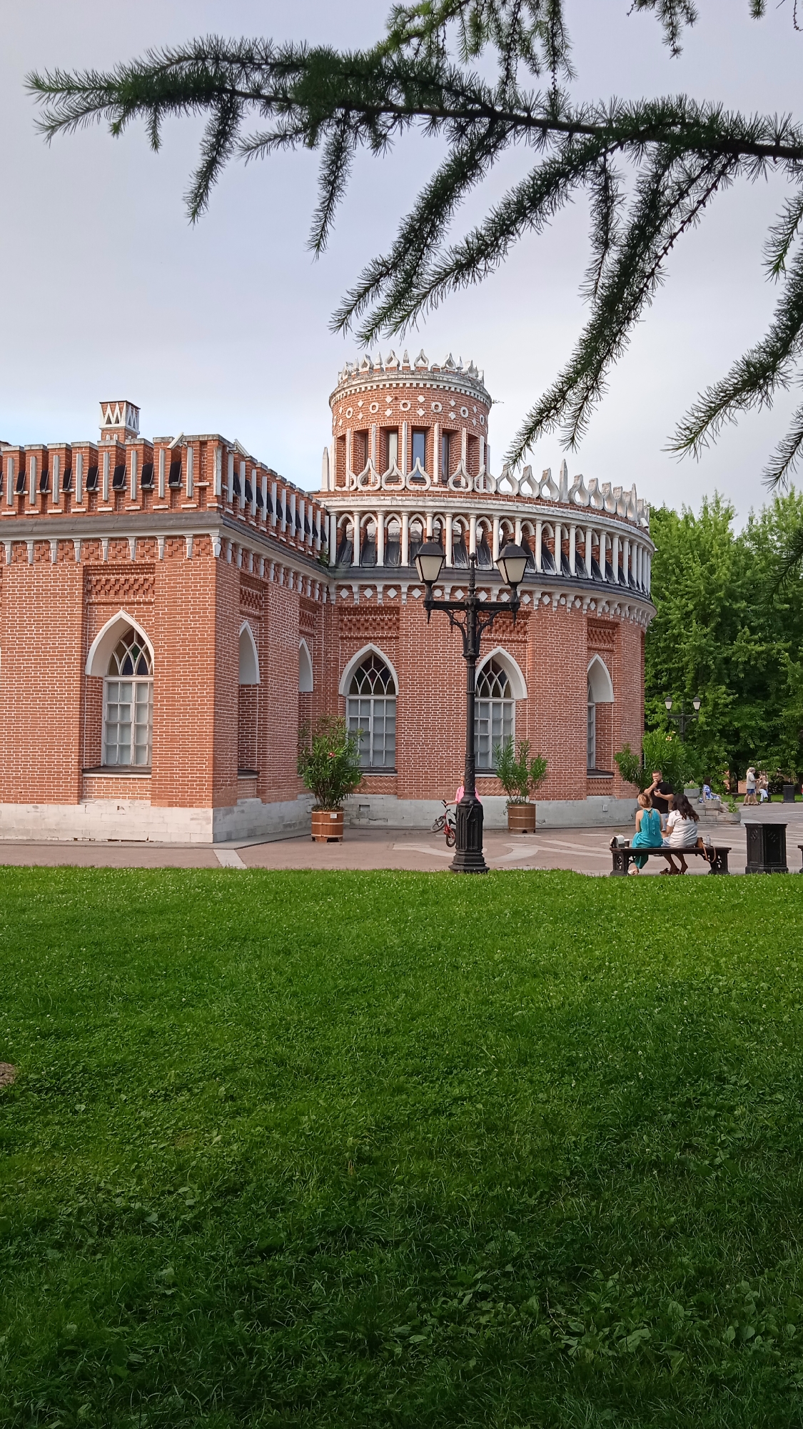 The image shows the main building of Tsaritsyno Museum-Reserve in Moscow, Russia. The building is made of red brick with white ornate detailing. It features a cylindrical tower and a long wing with arched windows. In front of the building, there is a green lawn, a lamppost, and a bench. Three people are sitting on the bench, and a bicycle is parked nearby. In the background, there are trees and other people walking. The sky is overcast.