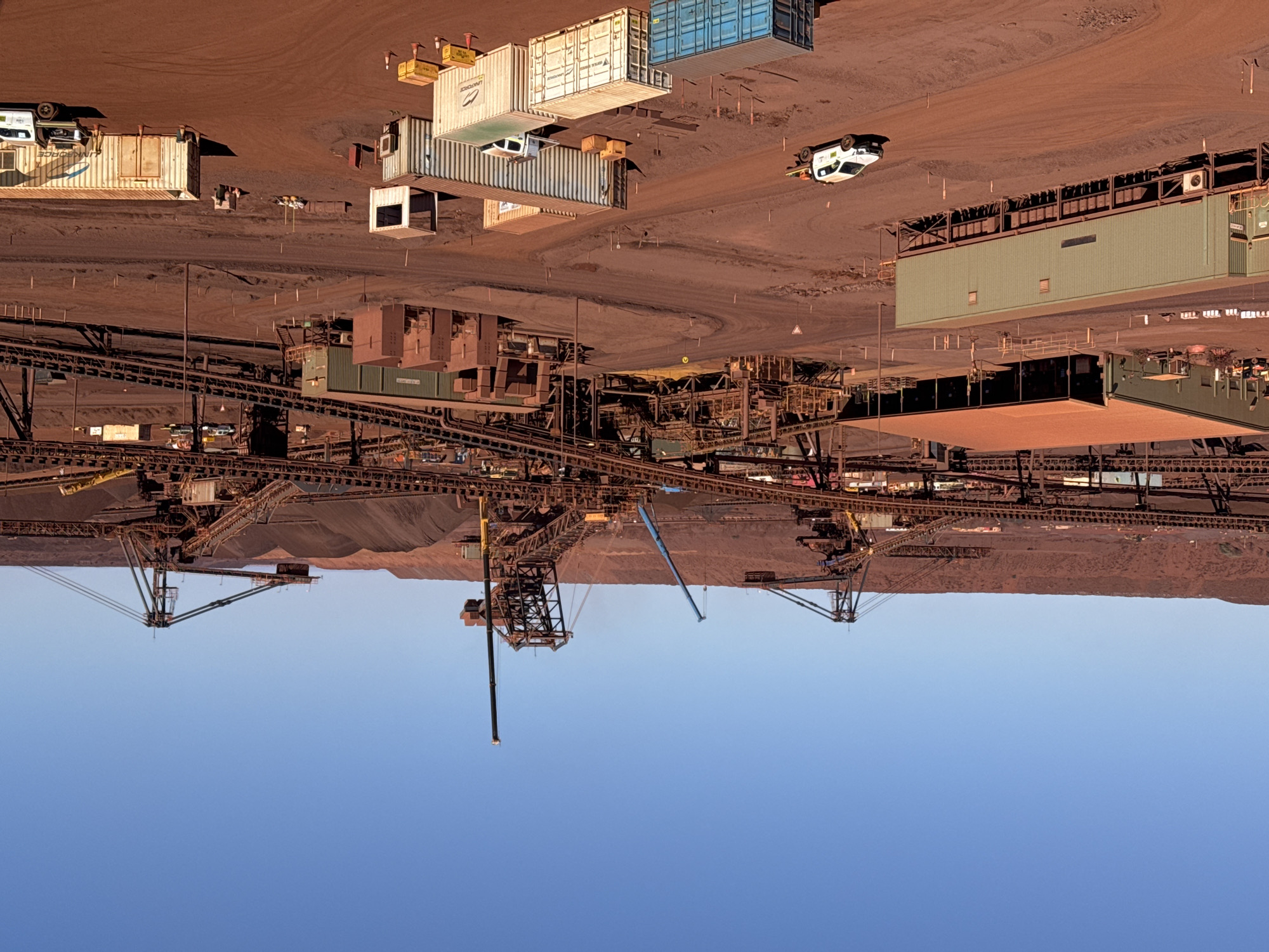 This is a wide-angle, high-angle shot of a large industrial mining operation in Newman, Australia. The foreground is dominated by a vast expanse of reddish-brown earth, characteristic of the iron ore-rich Pilbara region. Scattered across this terrain are various elements of the mining infrastructure.
In the lower left quadrant, a long, light green shipping container-style building is elevated on stilts. A white utility truck is parked on a dirt track in front of it, facing right. Further into the midground, several more shipping containers, in various colors including white, cream, and blue, are arranged in clusters. Some have branding visible, such as "UNI FORCE." Yellow cones and wooden crates are placed around some of the containers.
Behind the foreground structures, a complex network of industrial machinery is visible. Multiple conveyor belts, supported by sturdy metal frameworks, snake across the scene, transporting materials. Large, towering mining equipment, including what appear to be stackers and reclaimers with their distinctive boom arms, are positioned amidst piles of dark, processed ore. A blue crane is also visible in the mid-upper portion of the frame, likely involved in construction or maintenance.
In the background, a massive, arid landscape of rolling hills or man-made mounds of ore stretches towards the horizon. The sky above is a clear, bright blue, indicating a sunny day. The overall impression is one of heavy industry operating in a remote, arid environment. There are no discernible people in the image, but the presence of vehicles and operational machinery suggests ongoing activity.