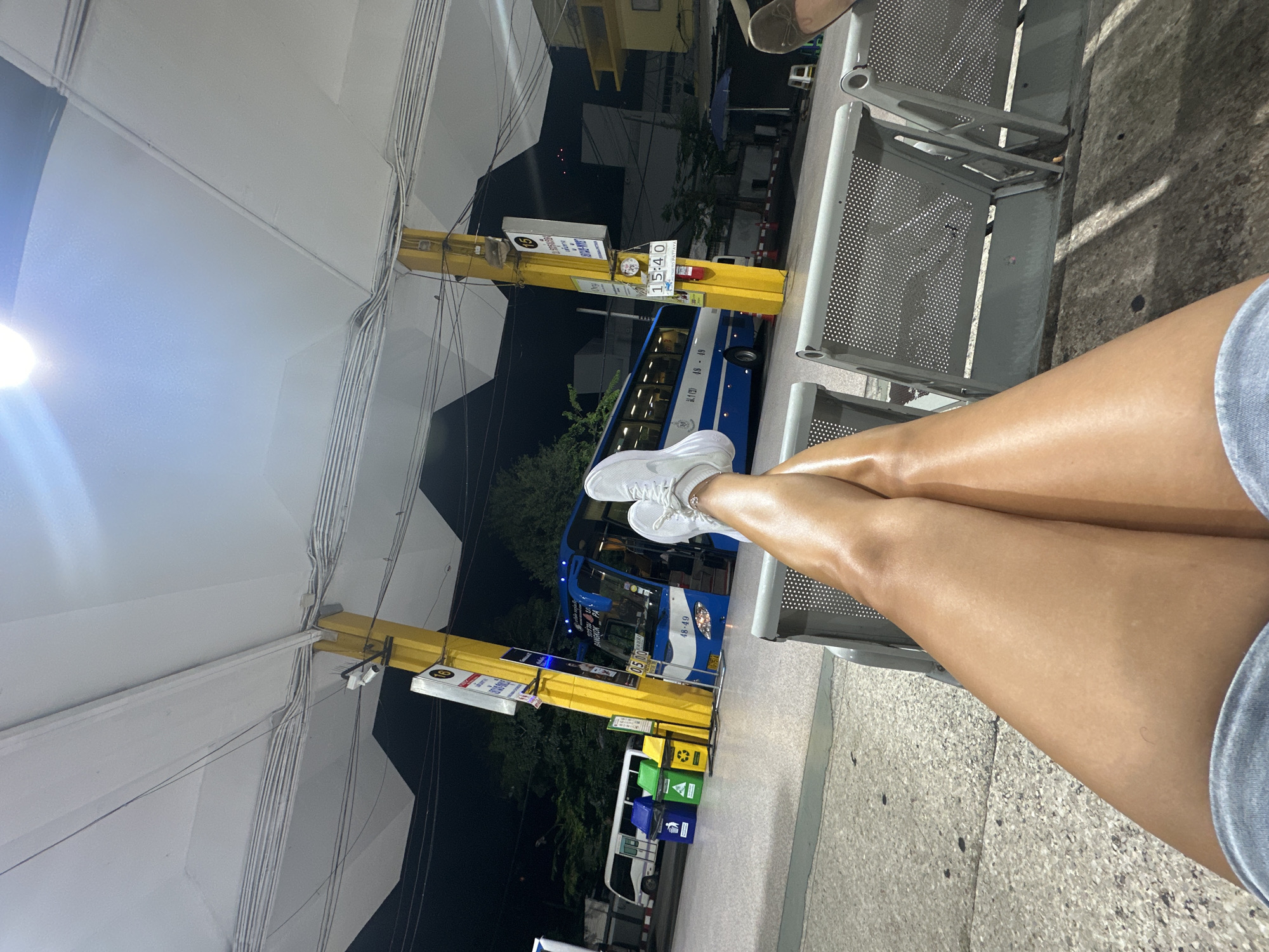 The image, taken at night, depicts a person sitting on a metal bench at a bus stop in Bangkok, Thailand. The person is wearing white sneakers. A blue bus is parked at the bus stop, which has a yellow sign displaying the time as 15:40. Multiple recycling bins are present near the bus stop. Overhead, there is a structure that appears to be the bus stop shelter, with a white ceiling and wires. The surrounding environment is dark, with visible trees and buildings.