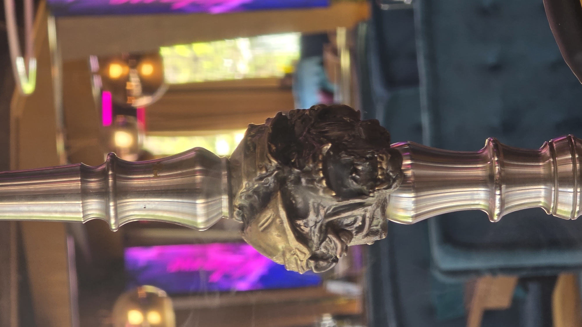 The image shows a decorative metal hookah with an ornate centerpiece, possibly a human or animal figure. The hookah is positioned horizontally against a blurred background, including lights, a wooden frame, and hints of outdoor greenery, possibly a window view. The foreground is slightly out of focus, with a hint of smoke emanating from the hookah. The hookah itself is a polished metal with detailed engravings. The location, Sofia, Bulgaria, is based on external knowledge and not visually identifiable from the image.