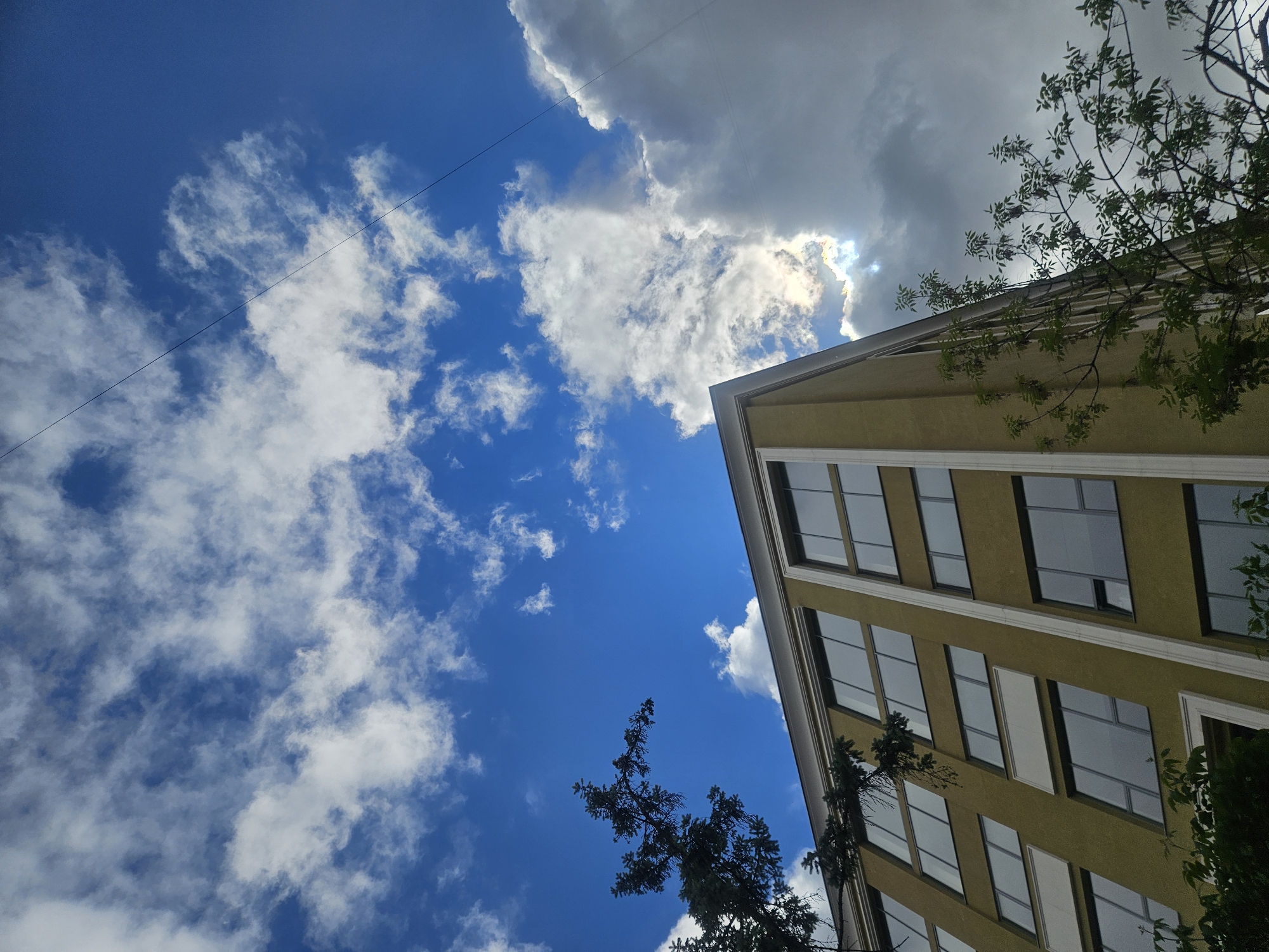 The image captures a low-angle perspective of a building juxtaposed against a dynamic sky. The building, located in Moscow, Russia, features a pale yellow exterior with multiple windows that reflect the sky and surroundings. It's a multi-story structure with a clean, modern design.
The sky dominates the frame, showcasing a mix of dark and light clouds scattered across a vivid blue expanse. A thin power line cuts across the sky, adding a sense of urban infrastructure.
Trees with green foliage are visible along the right side of the building and at the bottom, partially obscuring the lower levels. The overall scene conveys a sense of urban architecture intertwined with nature under a changeable sky.