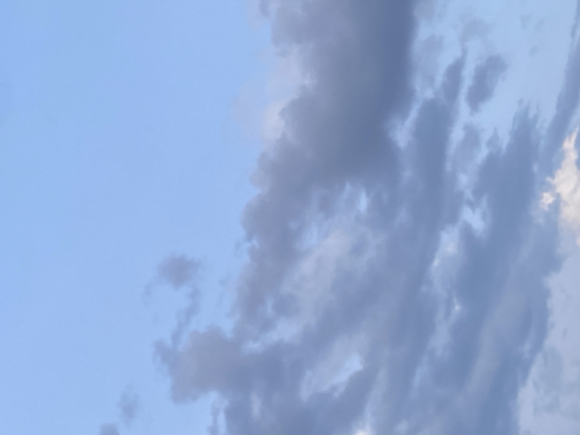 The image depicts a daytime sky in Tinaquillo, Venezuela. The left portion of the frame shows clear, light blue sky. The right side is filled with layered, textured clouds ranging in shades of gray and pale blue. The clouds appear somewhat dense, obscuring parts of the sky behind them. No people, objects, or actions are visible within the frame other than the natural environment of the sky and clouds.
