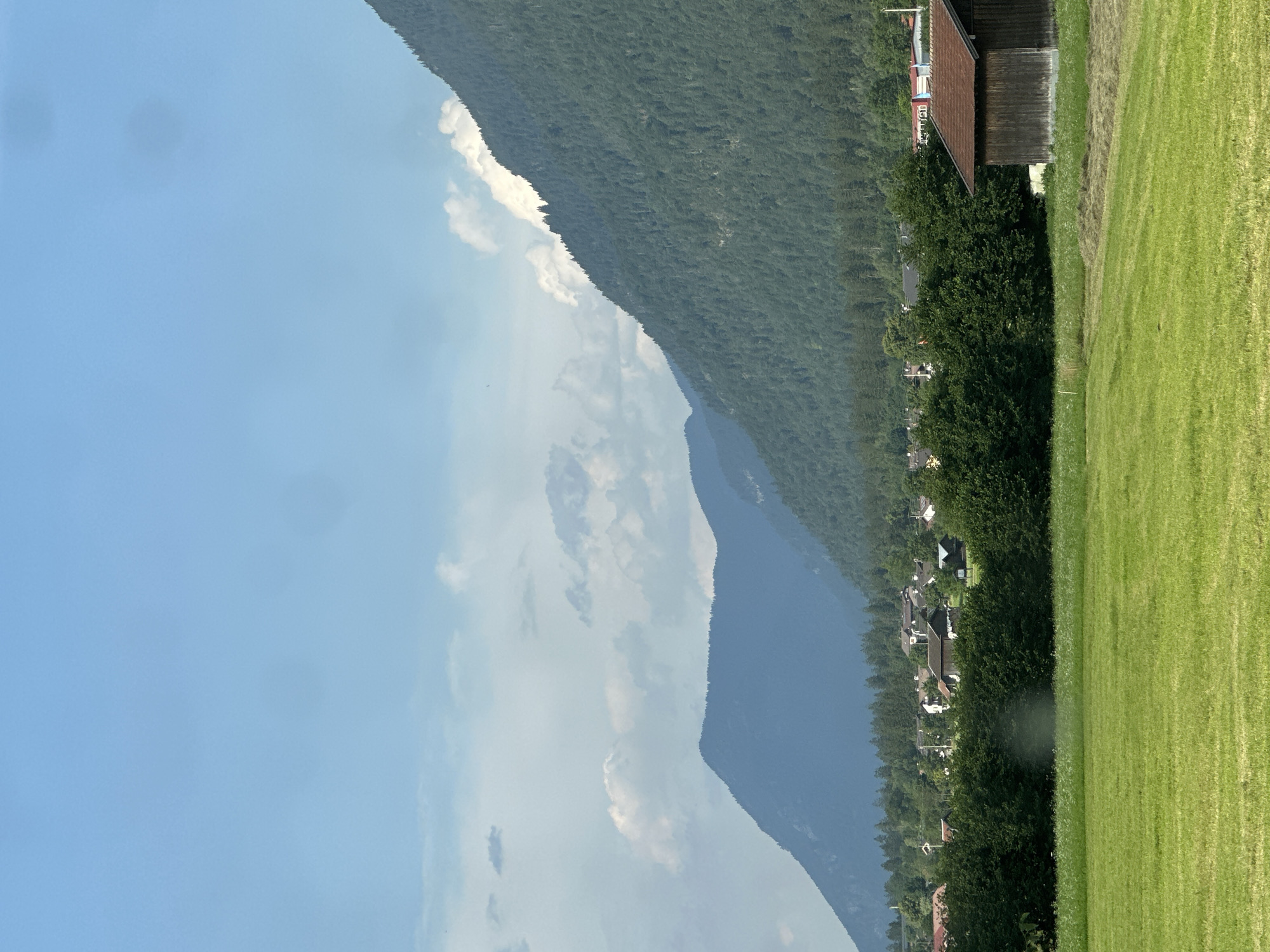 The image depicts a rural landscape in Garmisch-Partenkirchen, Germany. The scene is dominated by a large, tree-covered mountain on the left, with the sky and clouds to its left. A small village is nestled at the base of the mountain, surrounded by lush green trees. In the foreground, a neatly mown green field stretches to the right, with a wooden building and a red-roofed structure visible at the edge of the field. The overall impression is one of a serene, natural setting with a touch of human habitation. The time of day appears to be daytime, possibly late afternoon, given the lighting. The weather seems clear with scattered clouds. The image is oriented vertically, suggesting it might be a photograph taken with a phone camera in portrait mode. There is no visible text in the image.