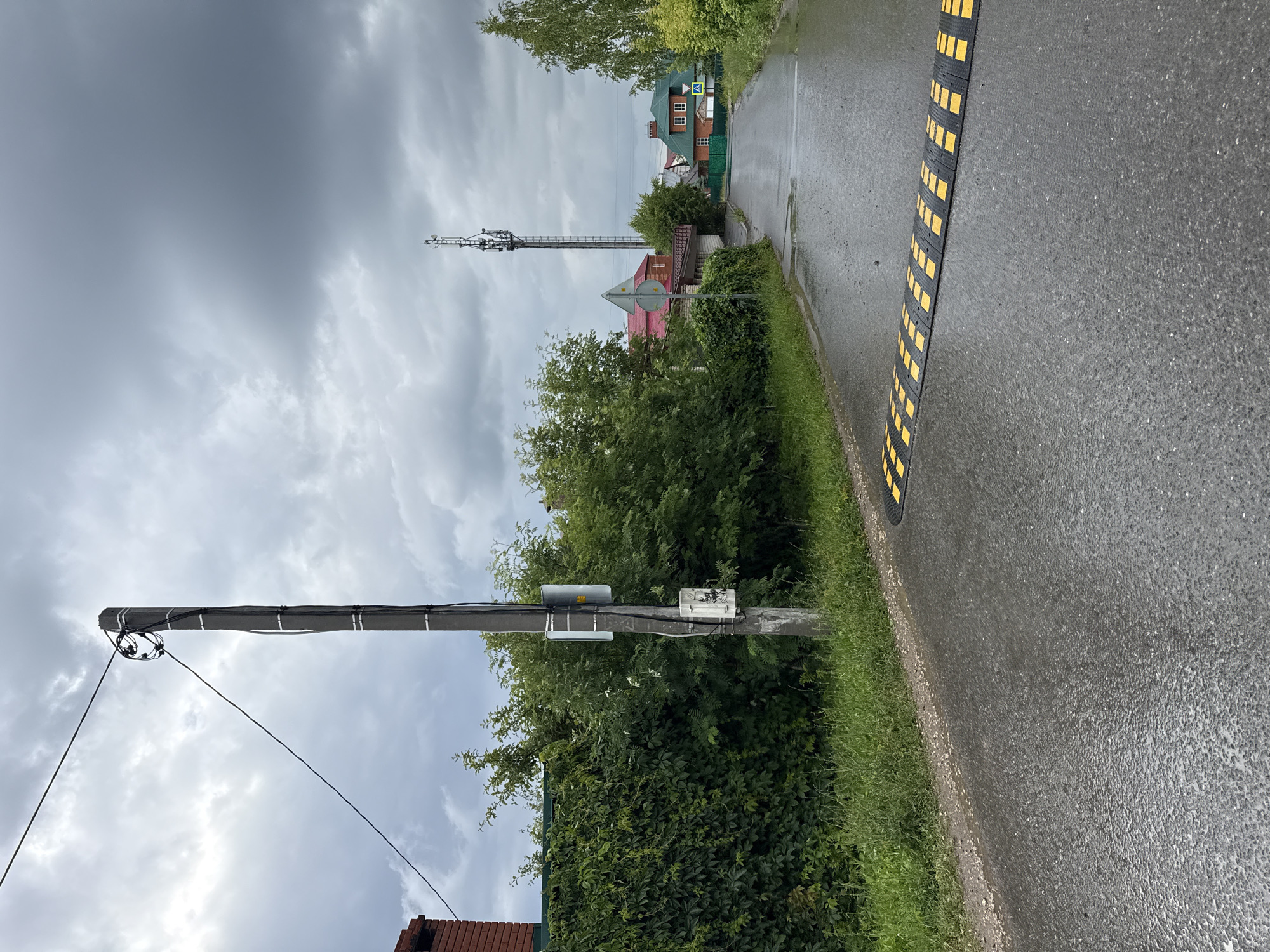 A wet, asphalt road curves slightly to the right, leading into a village under a cloudy sky. A black and yellow speed bump stretches across the road in the foreground. To the left, a tall, concrete utility pole stands next to a dense, green ivy-covered fence. Beyond the fence, lush green bushes and trees are visible, with a red brick chimney peeking out from behind the vegetation. In the background, several houses with red and green roofs dot the landscape. A tall telecommunications tower with multiple antennas rises above the buildings. A pedestrian crossing sign is visible on the right side of the road. The overall scene suggests a damp, overcast day in a suburban or rural area.