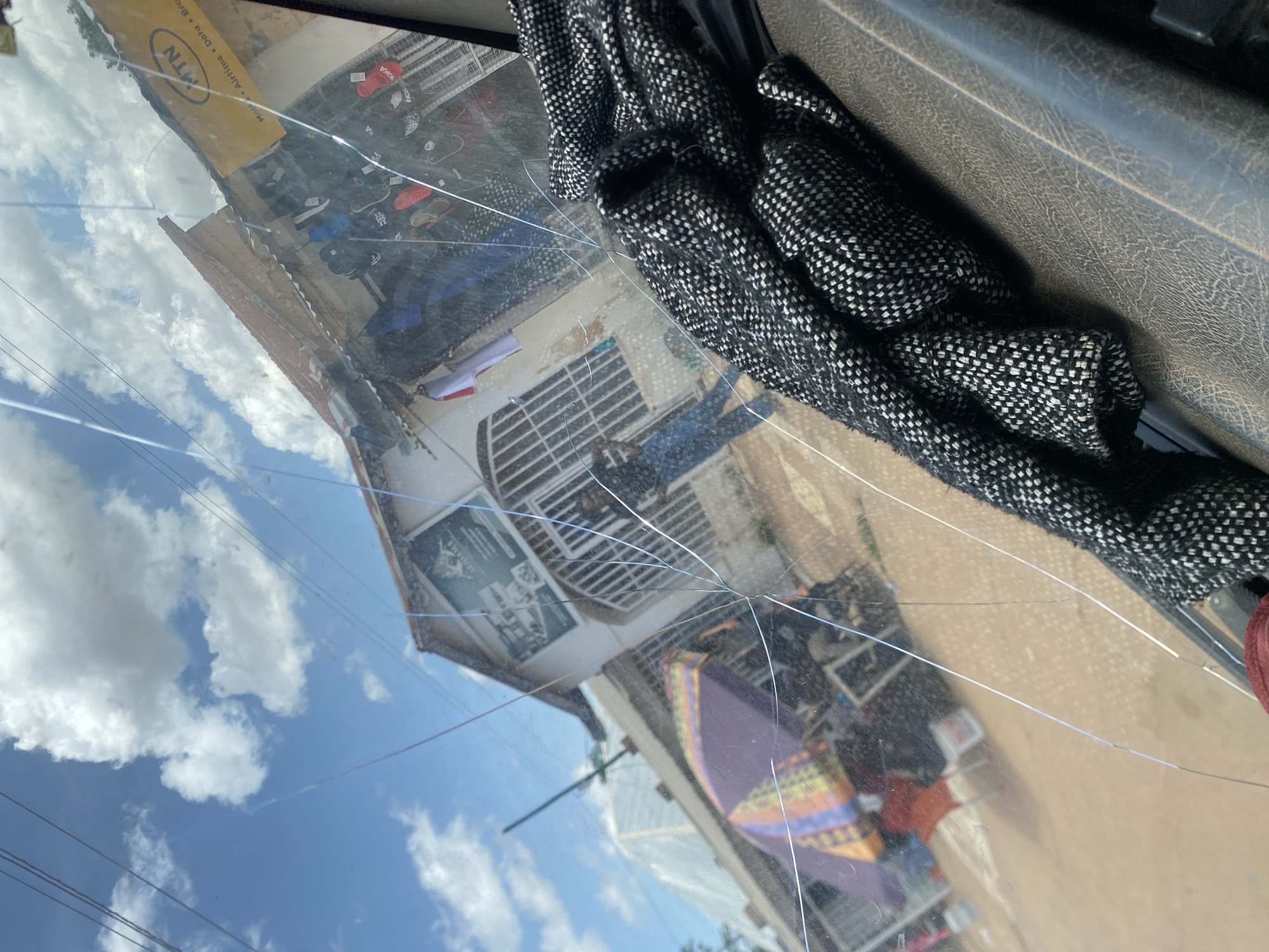 The image depicts a view through a cracked windshield of a street scene in Gura Top, Nigeria. The foreground is dominated by the cracked glass and a textured, dark-colored dashboard or interior surface, with a black and white patterned fabric draped over it.
In the mid-ground, the scene outside reveals a modest building with a sloped, reddish-brown roof. A sign with text and an emblem is visible above an entranceway. The building appears to be a shop with items displayed behind a metal gate or grill. Various merchandise, including what seem to be shoes and clothing items, are visible through the glass. A yellow MTN sign is also prominent on the building's facade. To the left, an umbrella with colorful stripes provides shade for a market stall. People are visible in the background, including one individual in the shop doorway wearing blue jeans and a dark shirt, and others near the market stall.
The environment is outdoors, with a bright blue sky filled with white, fluffy clouds. The overall impression is of a sunny day in a busy, potentially commercial area of a Nigerian city. The cracks in the windshield create a fragmented view of the external environment.