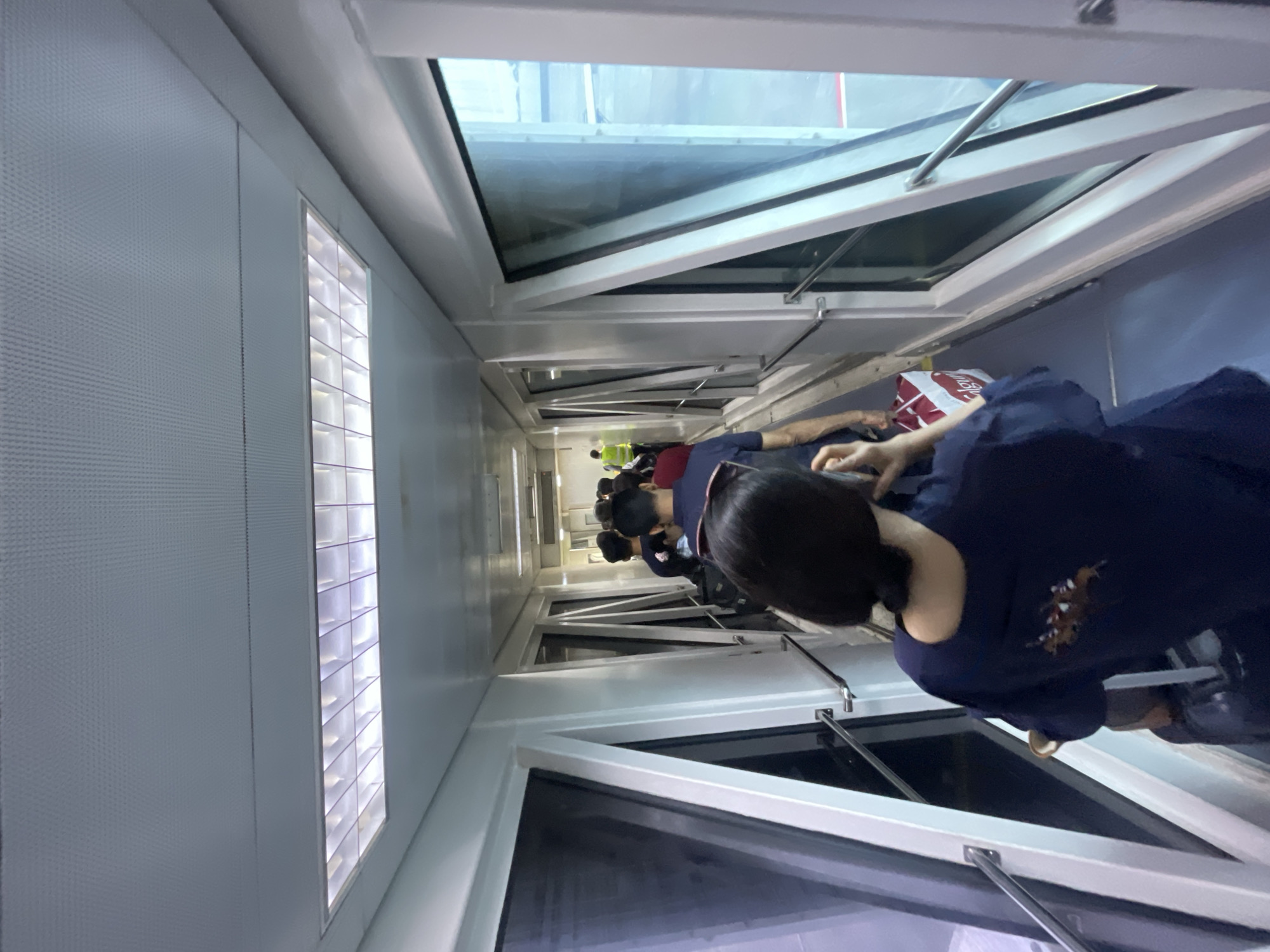 A group of people are walking through an airport jet bridge. The jet bridge is enclosed with large windows on the right side, offering a view of the tarmac and an airplane. The interior of the jet bridge is lit by fluorescent lights in the ceiling. The people are dressed casually and are carrying luggage, suggesting they are boarding or deplaning an aircraft. One person in the foreground is facing away from the camera, looking down at their phone. In the background, a person in a high-visibility vest is visible, possibly an airport worker. The overall scene depicts a common airport transit environment.