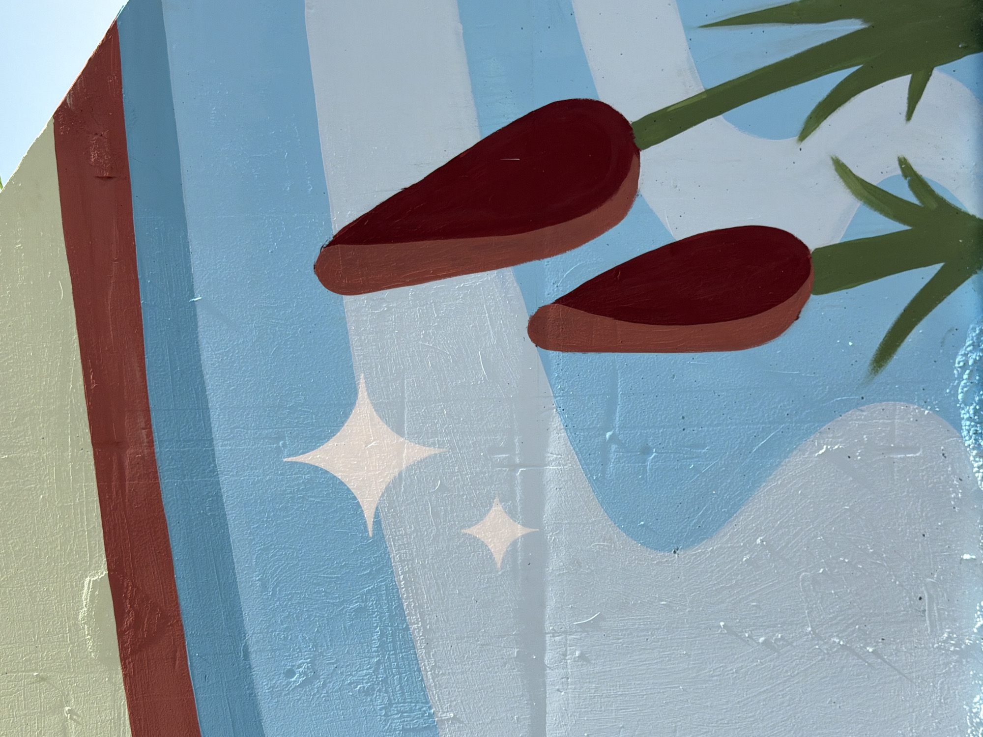 The image is a close-up of a mural on a wall, likely in Yekaterinburg, Russia, depicting a stylized landscape with abstract elements. The mural features bands of color: a light green band at the top, followed by a dark red band, and then a light blue band with white horizontal stripes. Below this, a lighter blue area creates a flowing, wavy shape that occupies the lower half of the image. Two abstract, dark red, bulbous shapes resembling cattails or stylized flowers are visible on the right side, emerging from green stems and blades of grass. Two white four-pointed star shapes, possibly representing sparkles or stars, are scattered in the lighter blue area. The wall itself has a textured surface, visible in the paint application and slight imperfections. The overall impression is a simple, graphic depiction of nature or a whimsical scene.