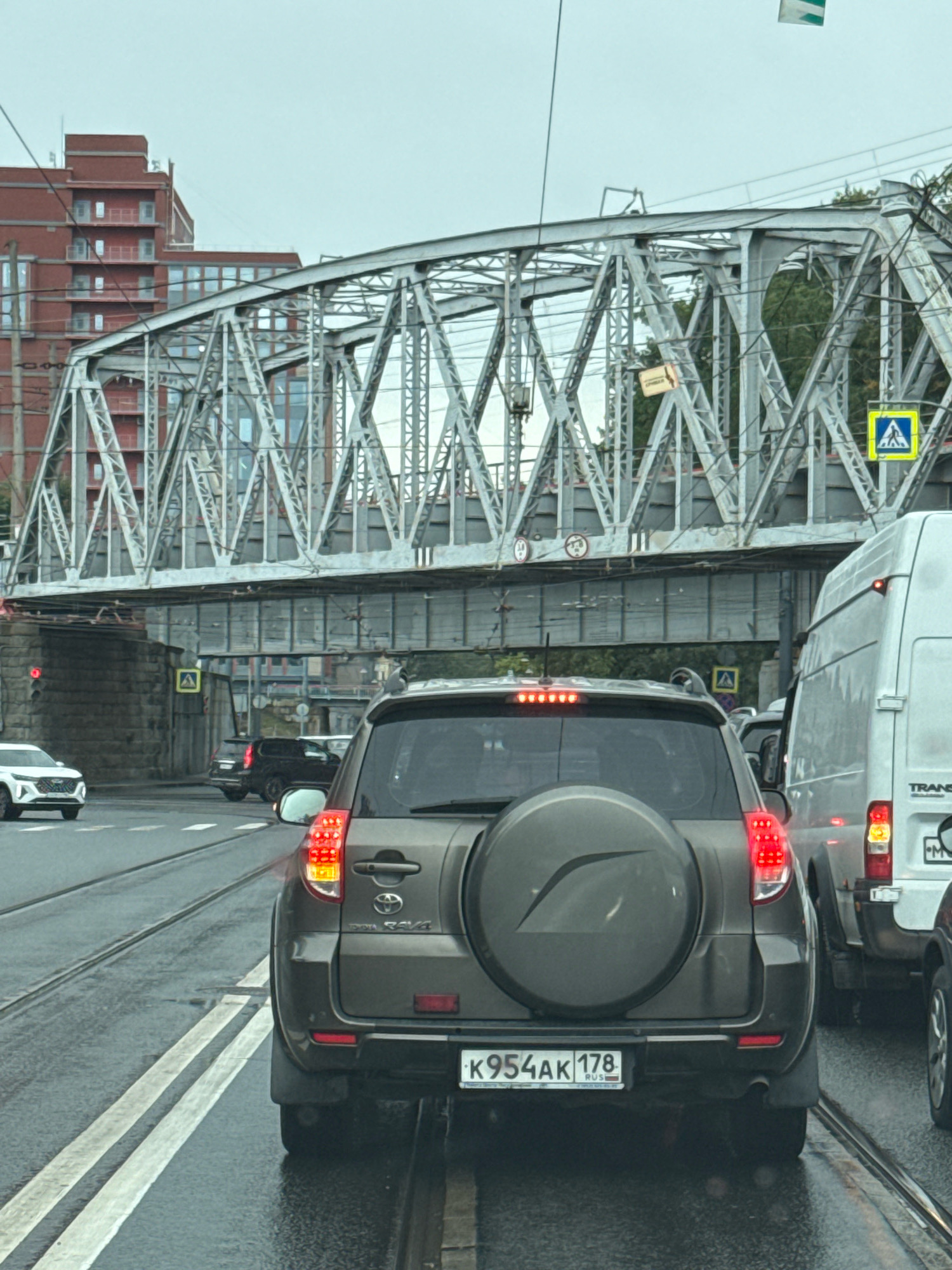 The image captures a traffic scene in Saint Petersburg, Russia, under an overcast sky. The foreground is dominated by the rear of a dark grey Toyota RAV4 SUV, identifiable by its spare tire cover with a stylized leaf emblem and its license plate, which reads "K954AK 178 RUS". The SUV's brake lights are illuminated, indicating it is stopped or slowing down in traffic. To the right of the RAV4 is a white van, also stopped.
In the mid-ground, other vehicles are visible, including a white sedan in the left lane and a dark SUV further ahead. The scene is set on a wet asphalt road with tram tracks running through it. The road infrastructure includes traffic lights, one of which is red, visible on a stone structure to the left.
Dominating the background is a large, intricate metal truss bridge, a characteristic feature of Saint Petersburg's urban landscape. The bridge spans across the road, with a complex network of girders and supports. Beyond the bridge, a modern red brick building with multiple balconies can be seen, suggesting a densely populated urban environment. Several power lines and streetlights are also present. Yellow pedestrian crossing signs are visible near the bridge supports. The overall atmosphere is one of urban congestion during a potentially rainy day, given the wet road surface.