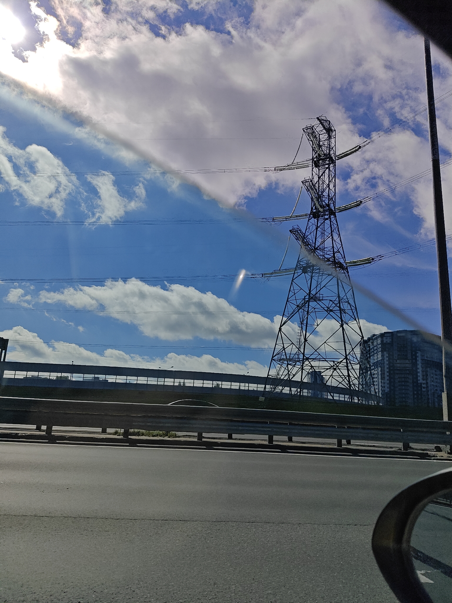 The image depicts a street-level view with a large electrical pylon as the dominant feature against a bright, partly cloudy sky. The pylon is a lattice-steel structure with multiple cross-arms supporting electrical cables. In the background, a modern, multi-story building is visible, suggesting an urban or suburban setting. A highway guardrail runs horizontally across the middle ground, separating the road from an elevated area with some greenery. The foreground shows a paved road, likely a highway, with the edge of a car's side mirror visible in the lower right corner. The lighting suggests it is daytime, with the sun casting bright rays through the clouds, creating lens flares and highlighting atmospheric haze. The overall scene conveys a sense of infrastructure and urban development within a bright, somewhat hazy daylight. The location is identified as Tarasovka, Russia.