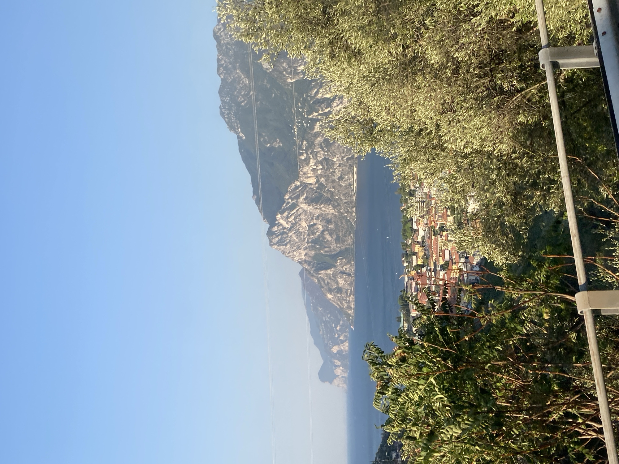 This is a photograph of a panoramic view of a town nestled along a body of water, likely a lake, with mountains in the background. The scene appears to be taken from a slightly elevated position, looking down towards the town and the water. The town's buildings have red-tiled roofs and are clustered together, suggesting a European setting. The water is a deep blue, and the mountains behind it are rugged and rocky, with patches of green vegetation. The sky is a clear, bright blue, indicating a sunny day. In the foreground, there are trees with green leaves, partially obscuring the view. A grey metal railing runs along the bottom of the image, suggesting it may have been taken from a balcony, overlook, or roadside viewpoint. The overall impression is one of a scenic landscape, characteristic of a lakeside town in a mountainous region. The query implies the location is Nago, Italy, which is consistent with the visual cues of Italian architecture and mountainous terrain often found in that region.