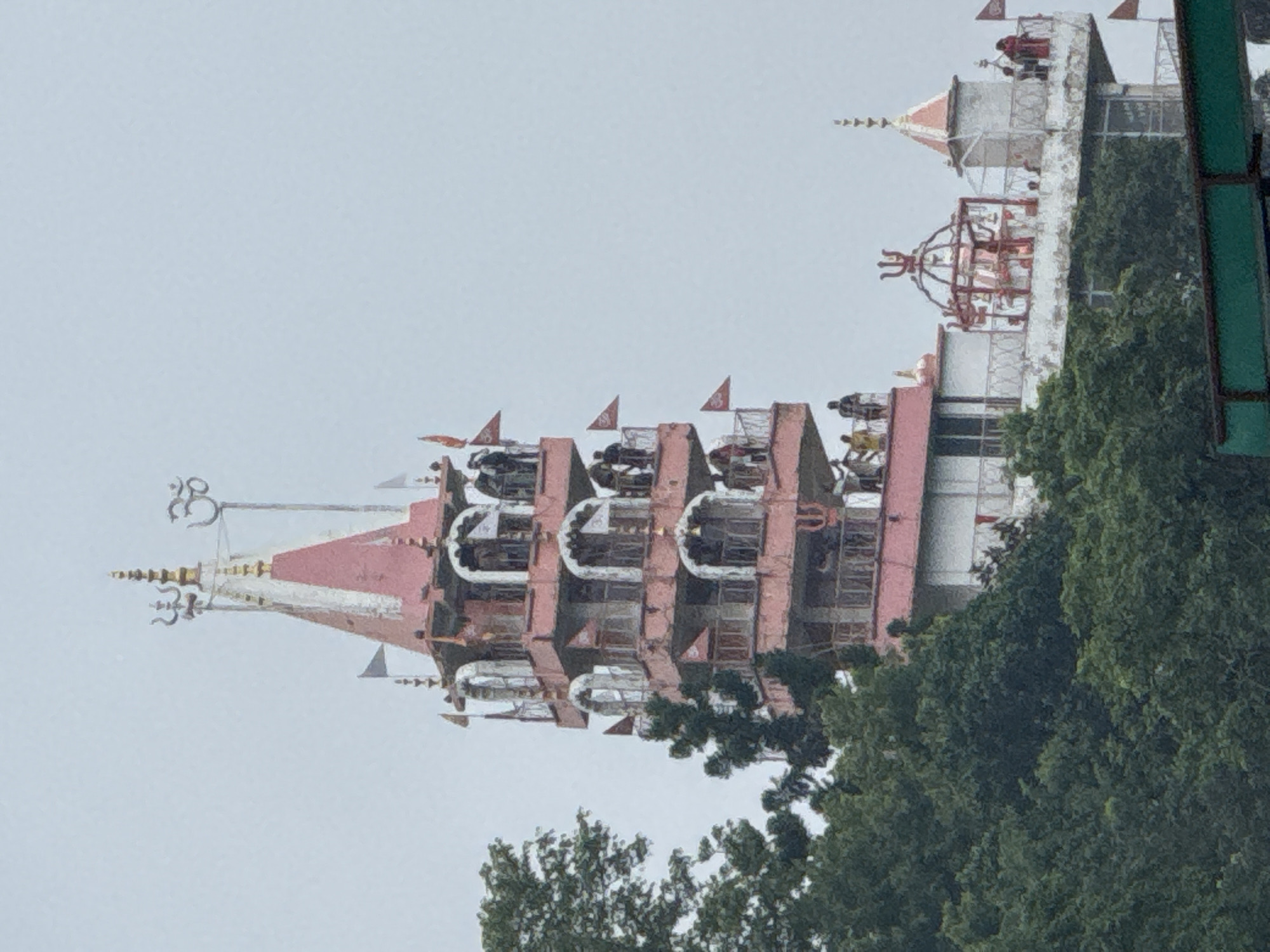 The image depicts a tall, multi-tiered pink temple structure situated amidst lush green trees in Rishikesh, India. The temple has several balconies and arches, with people visible on various levels, some standing and observing the surroundings. Small flags adorn the sides of the structure. At the very top of the temple, religious symbols including a trident and the Om symbol are visible. In the foreground, the tops of trees obscure the lower parts of the temple and the surrounding area. The sky is overcast, suggesting a cloudy day.