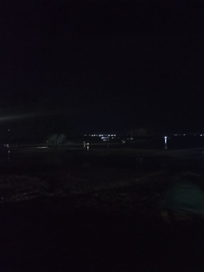 This media presents a dark, nocturnal landscape captured in Kecamatan Pujut, Indonesia. The overall scene is characterized by extreme low light, with artificial illumination being the primary visible elements.
In the foreground, the ground appears dark and indistinct, possibly a field or marshy area, with subtle variations in texture suggesting uneven terrain or wet patches. There are no discernible objects or subjects directly in the immediate foreground.
Moving towards the mid-ground, faint, dark outlines of what appear to be trees or shrubs can be glimpsed against the slightly brighter horizon, particularly in the central left area of the image. Reflections of distant lights are visible on what seems to be a body of water or extensively flooded ground, creating vertical streaks of light.
The background is dominated by a clear horizon line dotted with numerous small, bright, mostly white and yellow lights. These lights stretch horizontally across the width of the image, indicating a distant town, settlement, or perhaps an industrial area. A few individual, brighter light sources are also visible, casting distinct reflections on the water surface in the mid-ground. On the far left, a diffused, soft light source creates a subtle glow against the darkness, possibly from a nearby streetlamp or vehicle.
No specific activity or event is captured, and there are no visible people or discernible text. The image conveys a sense of quiet solitude, typical of a nighttime landscape view over a rural or semi-urban area. The atmosphere is calm, with the darkness punctuated only by the distant glow of human habitation.