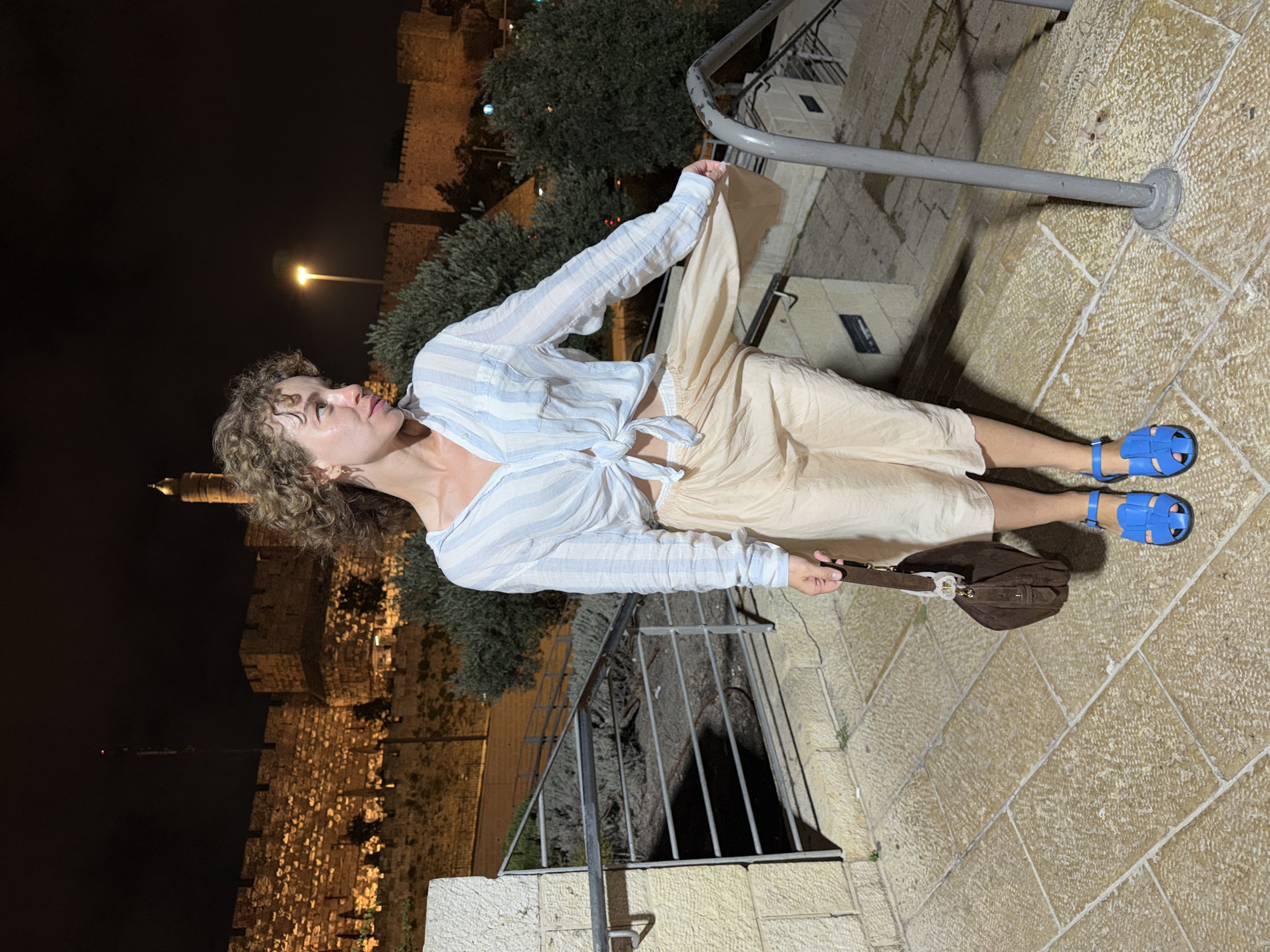 A woman with curly brown hair stands on a stone staircase at night, looking to her right. She is wearing a light blue and white striped, tied-up shirt and a light beige, flowy skirt. She holds a dark brown handbag in her right hand and wears bright blue sandals. In the background, a stone wall with glowing yellow lights stretches across the scene, with a tall tower visible on the right. Trees and other structures are dimly lit behind the wall. The sky is dark, with a single bright streetlamp visible.