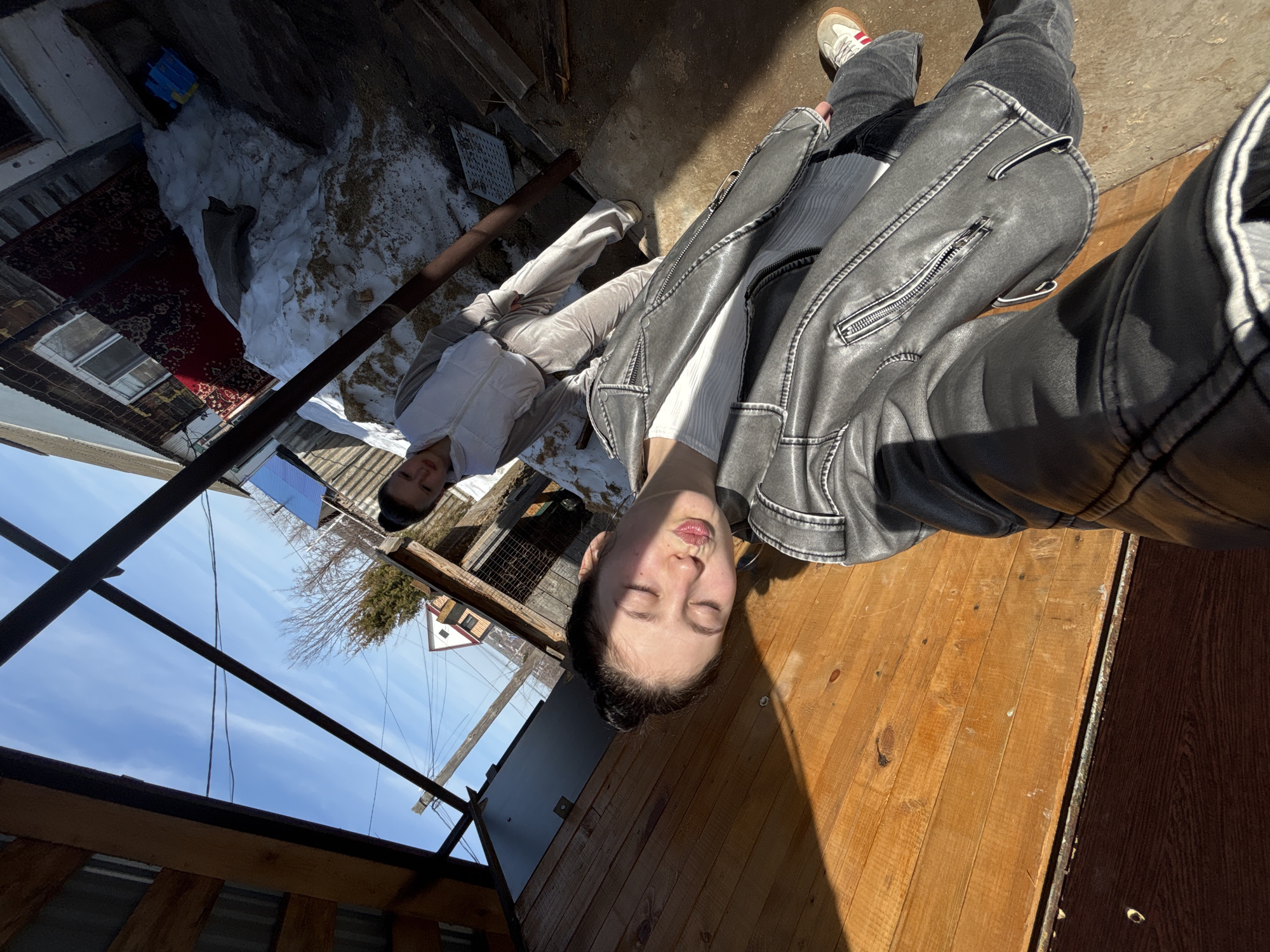 Two individuals are visible in an outdoor setting in Фоки, Russia. One is prone on wooden planks, clad in a white top and grey leather jacket, their eyes closed. The second person, wearing a white tracksuit, is lying in an area with lingering snow, situated beneath a metal support structure and a partial view of a building exterior. The immediate environment includes wooden decking, patches of snow, and a rug partially visible in the upper left. The sky is clear and blue.