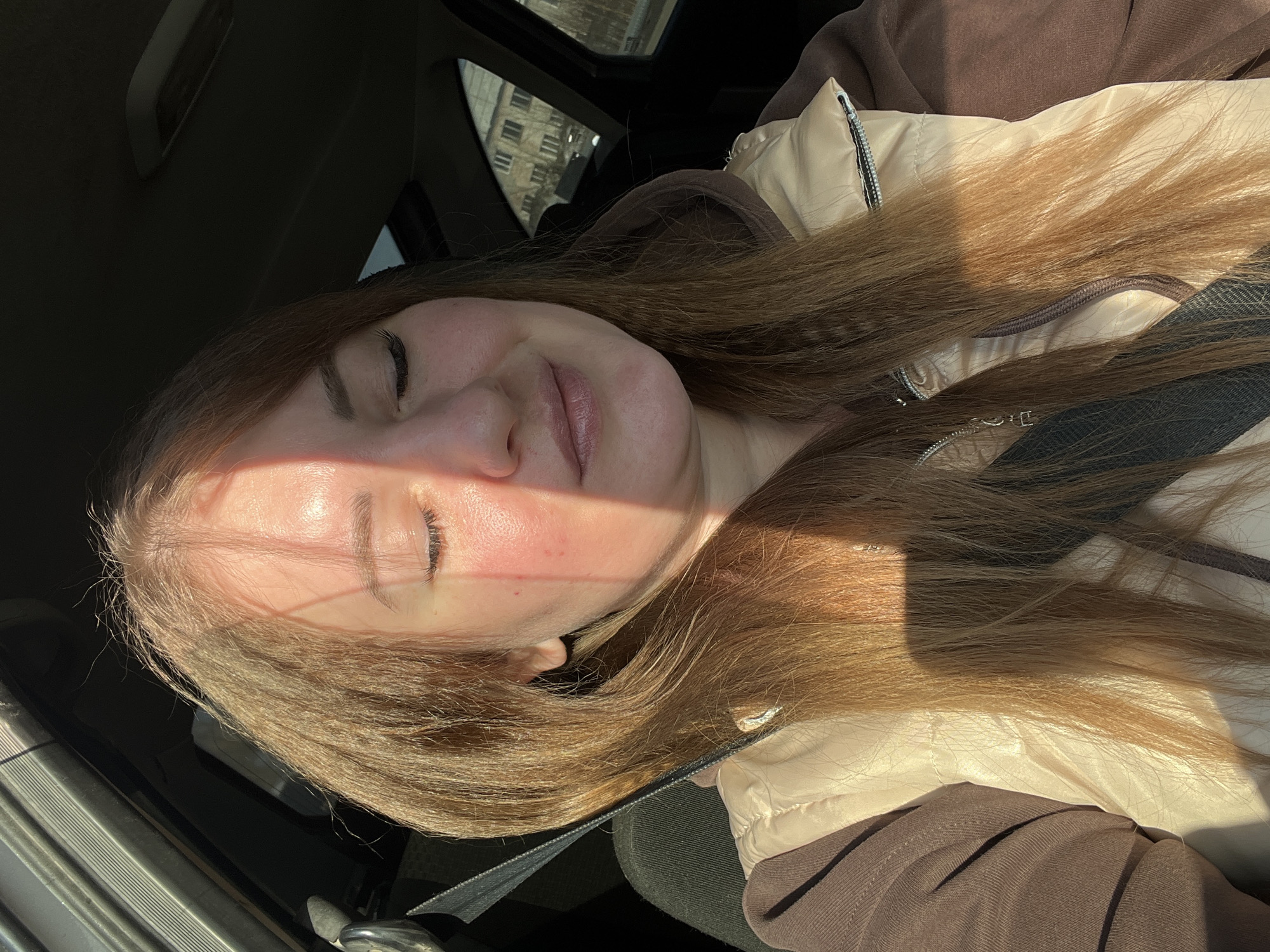 A person is sitting in a car. The person is wearing a seatbelt and a jacket and has brown hair. The buildings outside are probably in Chaikovsky, Russia. The person's eyes are closed. The sun is shining on her face.