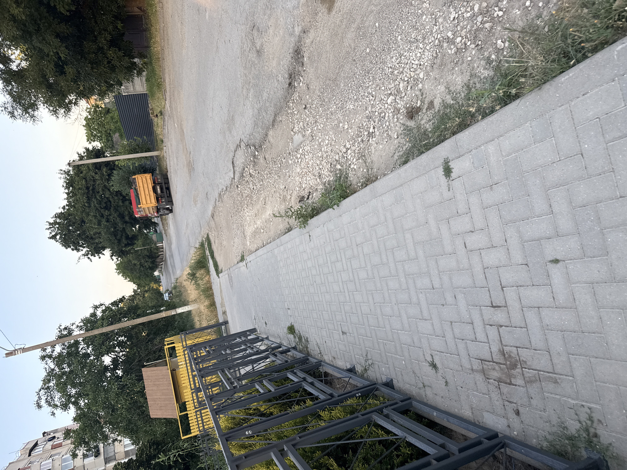 The image depicts a street scene in Cahul, Moldova, likely during the late afternoon or early evening given the soft lighting. A paved sidewalk runs along the left side of the frame, bordered by a dark metal fence. The sidewalk is constructed of light gray rectangular pavers laid in a herringbone pattern. Adjacent to the sidewalk, on the right, is a gravelly roadside with sparse vegetation and some debris.
The street itself appears to be asphalt, with visible signs of wear and tear, including potholes and uneven patches. A yellow and orange dump truck is parked on the right side of the street, facing away from the camera. In the background, various trees and buildings are visible, including a multi-story apartment building on the far left. A utility pole stands near the center of the image. The sky is a pale blue, indicating a clear day. The overall impression is that of a residential or semi-industrial street in a moderately developed urban area.