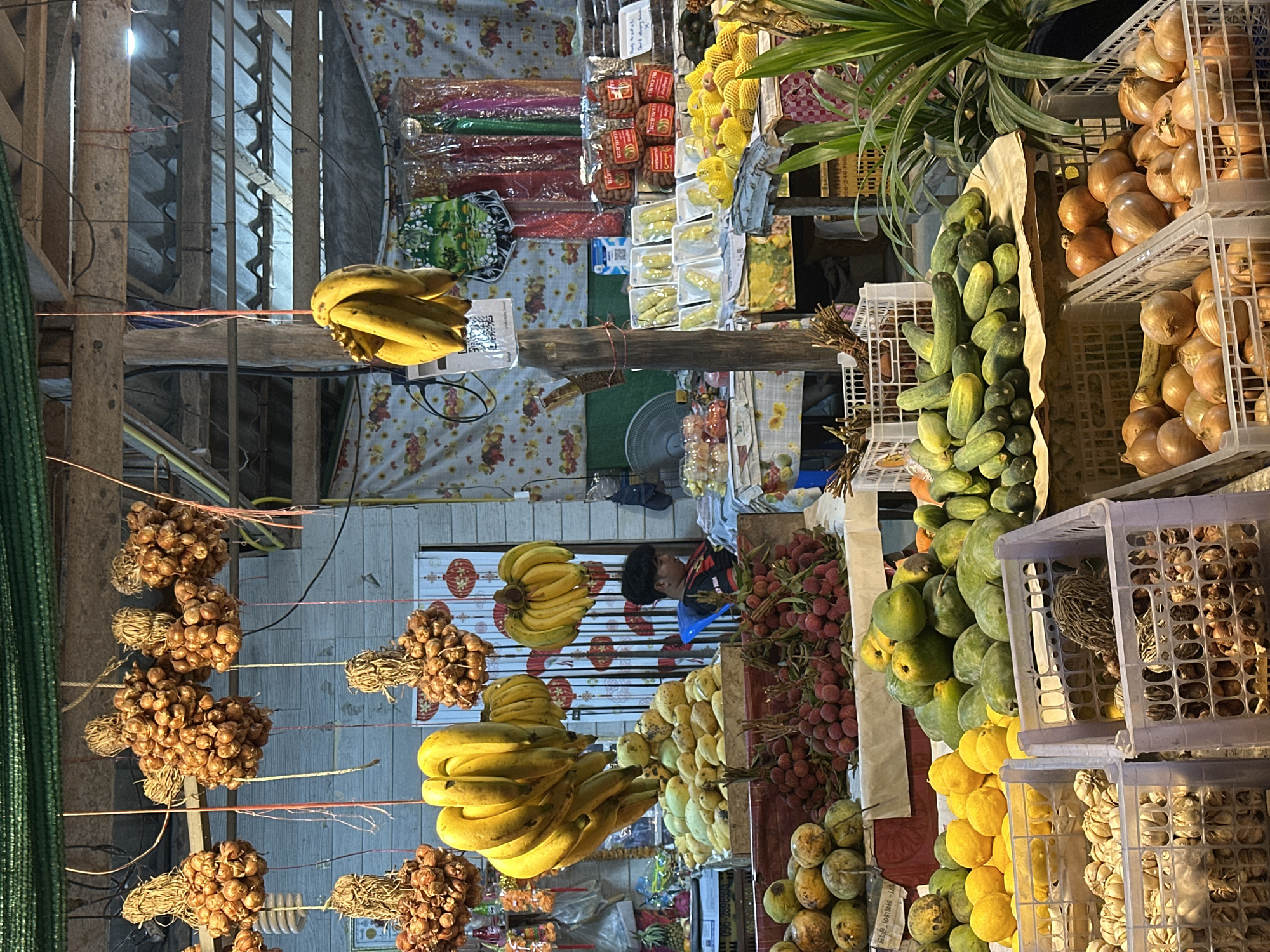 This image depicts an outdoor market scene in ตำบลเกาะพะงัน, Thailand, likely a fruit and vegetable stand. Bunches of dried garlic or onions hang from overhead structures, alongside clusters of ripe yellow bananas. Below, various fruits and vegetables are displayed. In the foreground, plastic crates hold onions and what appear to be garlic bulbs. A tiered display features yellow citrus fruits, mangoes, red lychees, and green cucumbers. Further back, more produce is visible, including what seem to be durian fruits packaged in plastic. A young person wearing a black t-shirt is seated on a blue plastic chair, looking down at a table. The market stall is covered by a wooden roof structure with corrugated roofing. Colorful fabrics with floral patterns serve as backdrops and coverings. The overall impression is a bustling, vibrant market environment.