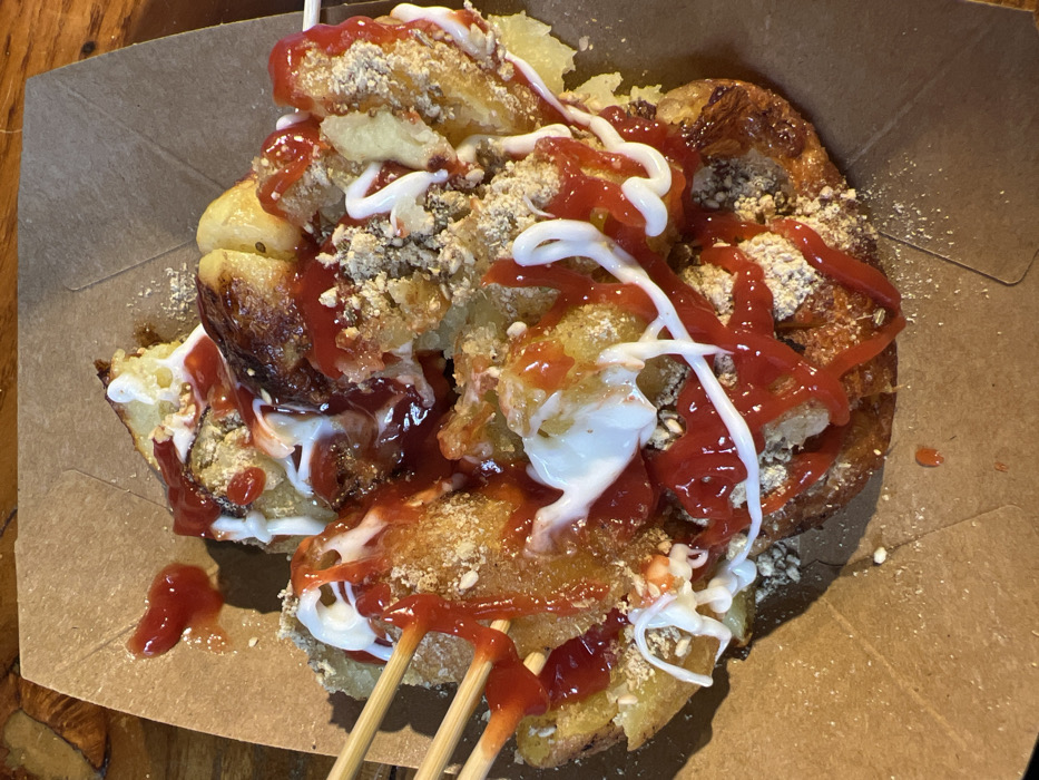 A close-up shows a serving of street food in a cardboard container. The dish features deep-fried potato pieces, drizzled generously with red ketchup and white mayonnaise. A scattering of crumbled, biscuit-like toppings adds texture. Two wooden sticks are partly visible, suggesting the dish is meant to be eaten with them. The food appears freshly prepared and served.