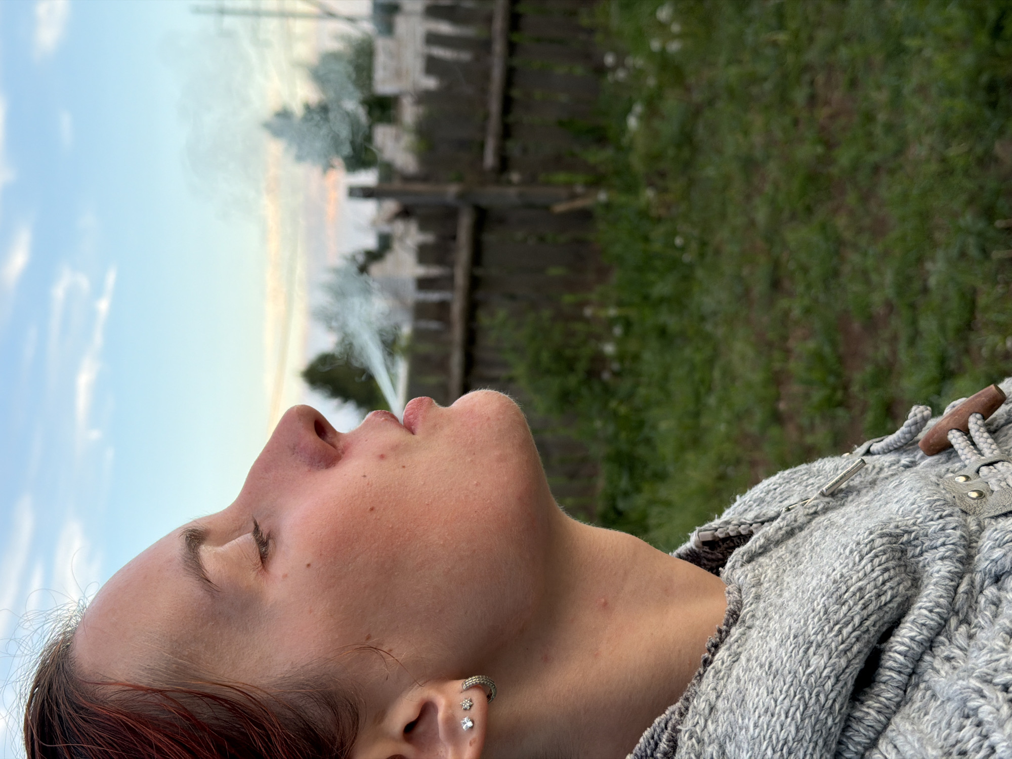 A medium shot, taken outdoors, depicts a person with fair skin, reddish-brown hair, and multiple ear piercings, tilted back with closed eyes and smoke exiting their mouth, presumably exhaling. The person is wearing a thick, grey, knitted sweater with toggle-style closures. The background consists of a blue sky with hints of orange and a wooden fence partially obscured by green foliage. The location cannot be determined as Chaikovsky, Russia based on the visual information alone.