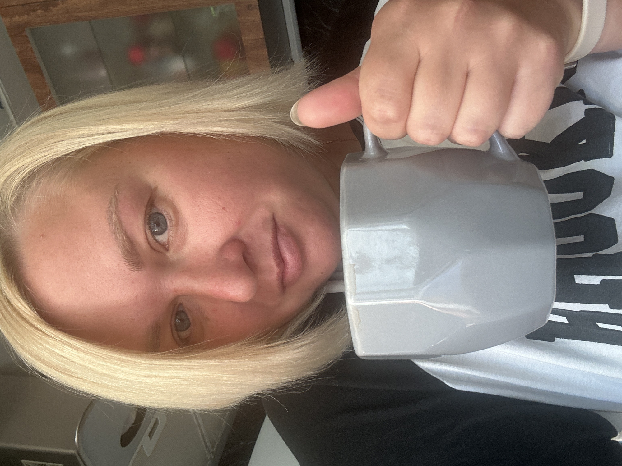 A blonde woman is holding a grey mug in her right hand, giving a thumbs-up with her left. She is wearing a black and white shirt with text on it. The background shows a kitchen cabinet and a light-colored exhaust hood. The image appears to be a selfie taken indoors. The location of Chaikovsky, Russia, is not discernible from the image.