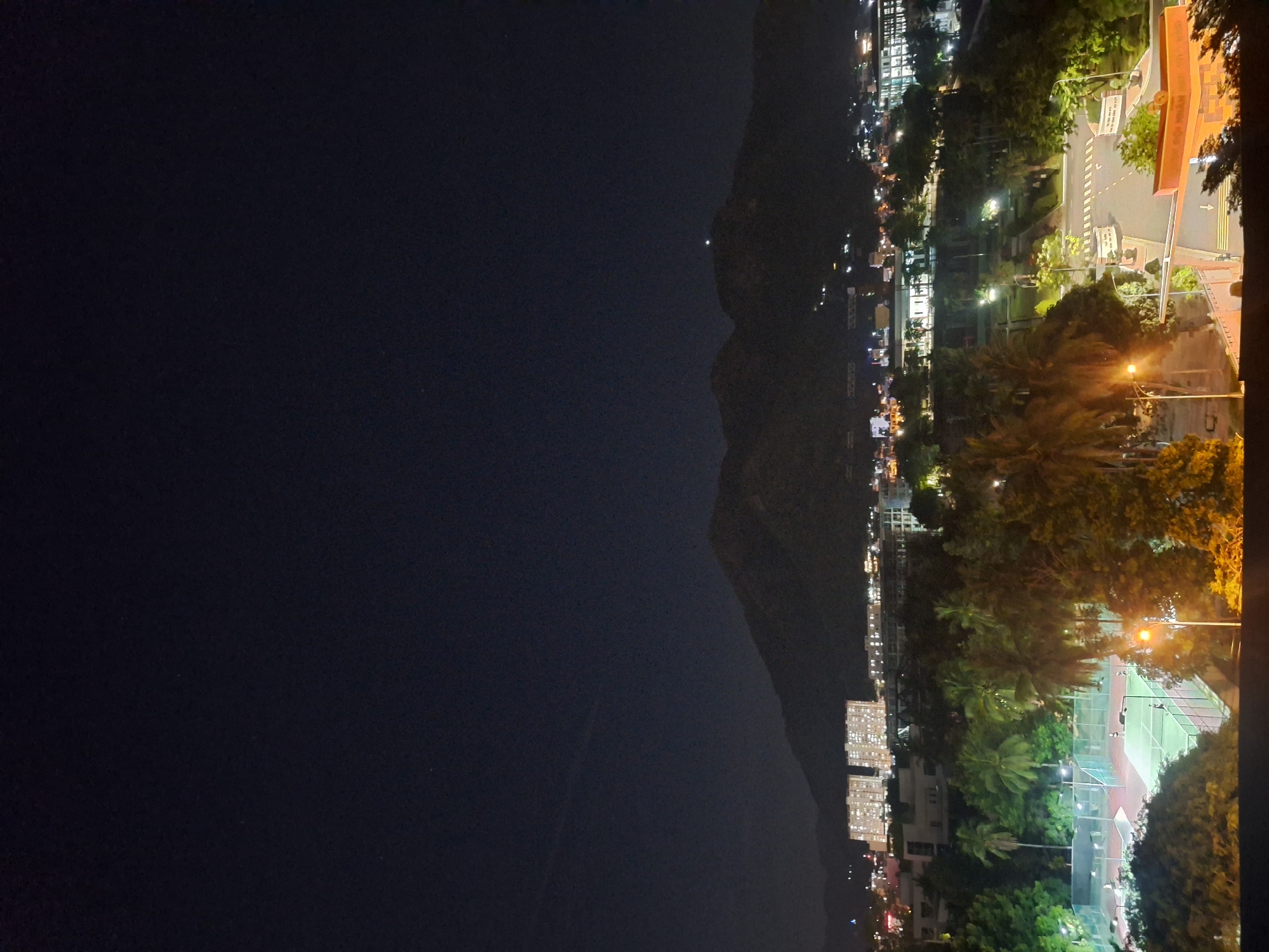 This image captures a nighttime view of a city landscape, likely Nha Trang, Vietnam. The foreground features a tennis court illuminated by artificial lighting, with several individuals engaged in playing tennis. Beyond the court, lush green trees, including palm trees, line the area. In the midground, illuminated buildings and a road system are visible, suggesting an urban environment. In the background, a silhouetted mountain range rises against the dark night sky. The overall scene depicts a well-lit recreational area within a city, bordering a natural mountainous backdrop.