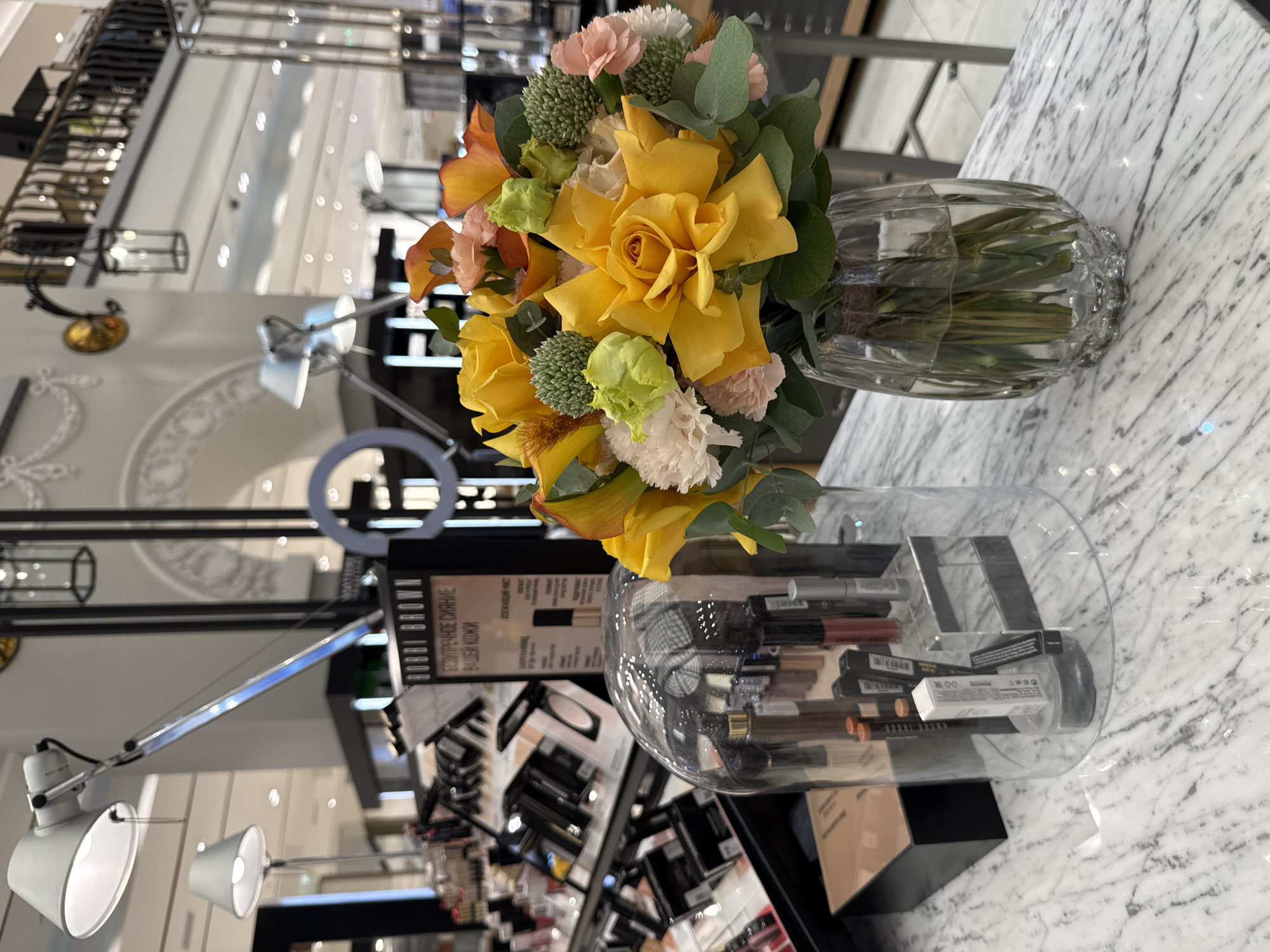This media file shows a close-up view of a marble countertop in what appears to be a retail setting, possibly a department store or a cosmetics shop. On the countertop, there is a glass vase filled with a colorful bouquet of flowers, predominantly yellow roses, with hints of pink, orange, and green accents. The flowers are lush and vibrant. To the right of the vase, a large, clear glass bowl is filled with various makeup products, primarily lipsticks and possibly other lip products, arranged in an organized manner. Some of these products are in their original packaging, while others are displayed as individual items.
Behind the countertop, a retail display area is visible. Several lamps, one with a white lampshade and another with a blue light, are positioned on the counter, suggesting a well-lit environment for product demonstration or display. In the background, there are shelves filled with more products, and the overall impression is of a clean, modern, and well-stocked retail space. The reflections in the background suggest a large, open area, possibly with a high ceiling and bright lighting, consistent with a retail store environment.
One visible text element is on a product in the background, which appears to be a Bobbi Brown cosmetic product. The text is in Russian and reads "БЕЗУПРЕЧНОЕ СИЯНИЕ ВАШЕЙ КОЖИ" which translates to "Flawless radiance for your skin". The overall mood is neutral and focuses on the presented items. The lighting suggests it is daytime, or a well-lit interior space. The location context indicates this is in Saint Petersburg, Russia.