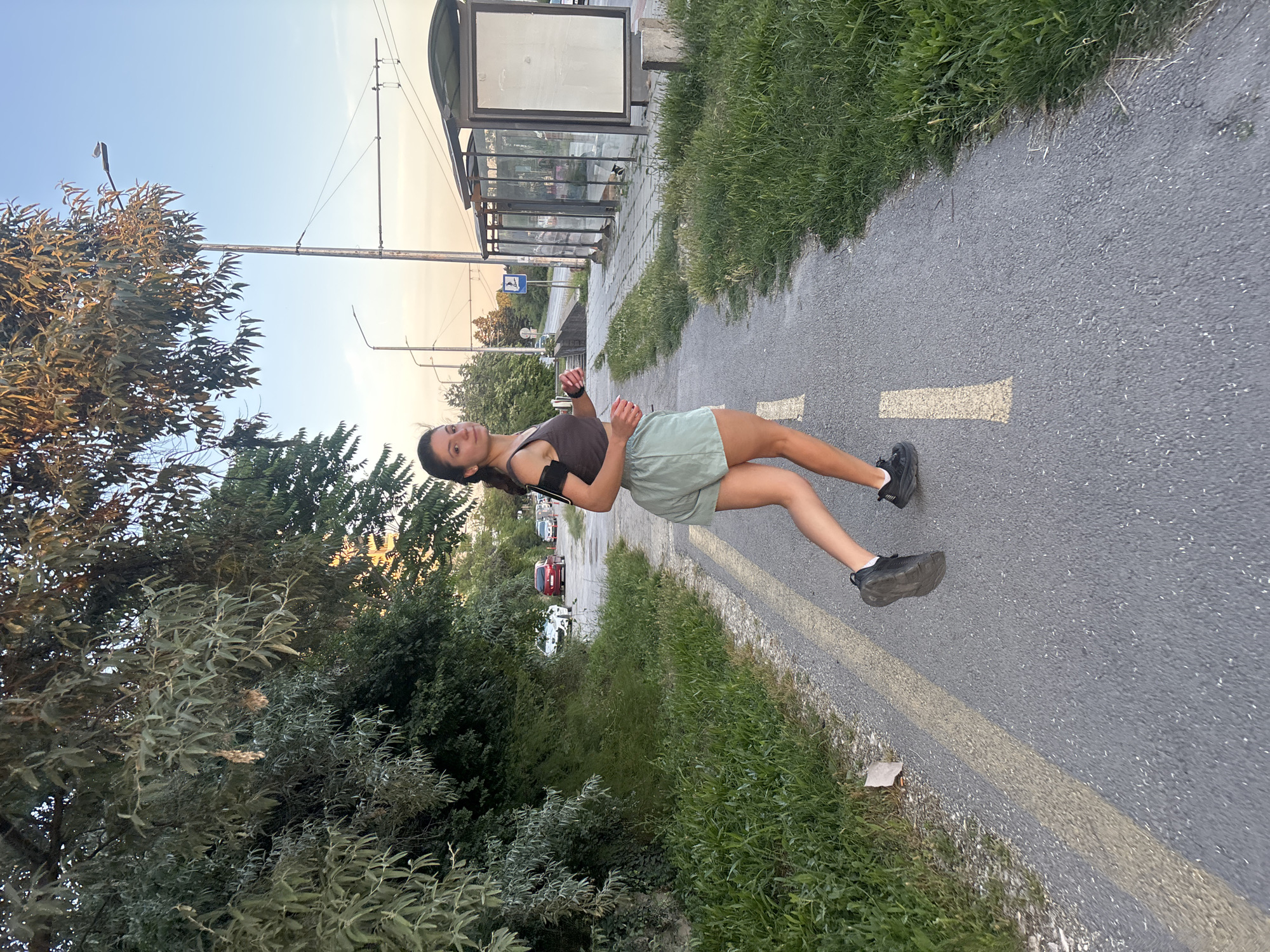 A young woman is jogging on a paved path with yellow lane markings. She is wearing a brown crop top, light green shorts, and black running shoes. An arm band is visible on her left arm. The path is bordered by a grassy area on the left and a sidewalk with a bus stop shelter on the right. Beyond the sidewalk, there are streetlights, traffic signals, and a road with cars. Lush green trees and foliage are present on the left side of the path, and some are visible in the background. The sky is clear with a gradient of blue and hints of orange, suggesting it is either early morning or late evening.
This scene appears to be located along the coastal road of Varna, Bulgaria, likely in the vicinity of the Sea Garden, a popular recreational area. The presence of the bus stop, streetlights, and the type of vegetation are characteristic of this seaside city's urban environment.