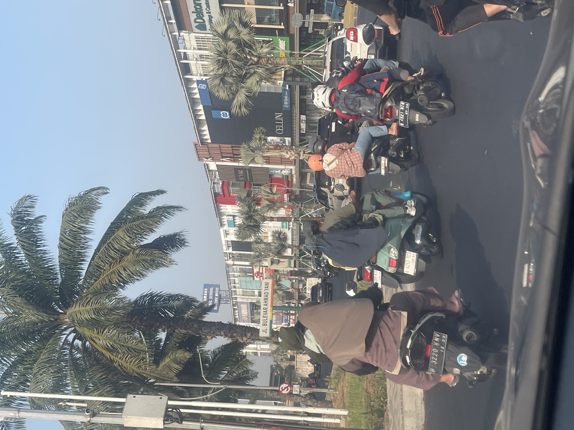 This is a street-level, eye-level shot of traffic on a sunny day in Bekasi, Indonesia. The scene depicts a moderately busy road with several motorcycles, a few cars, and a white delivery van. The foreground is dominated by the rear of a black motorcycle with a license plate reading "B 3220 KNV." The rider is wearing a brown tunic and a grey head covering. To the left, another motorcycle carries two passengers, one with a grey hoodie and the other with a light brown head covering.
Further back, a green scooter is visible, and to its right, a motorcycle with an orange helmet and a plaid shirt rider is seen. In the background, a white car and several other vehicles are present. The backdrop consists of a row of buildings with a large sign bearing "JL. BULEVAR AHMAD YANI." The buildings have various shop fronts, including "YAMAHA MUSIC SCHOOL," "CELLA," and "SUBWAY." Lush palm trees are scattered throughout the scene, with a particularly large one to the left. The sky is clear and blue.