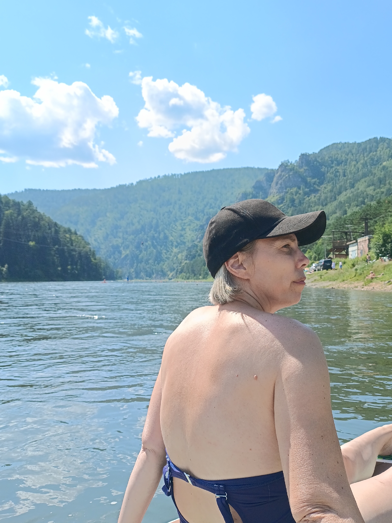 An overhead medium shot captures a woman from the shoulders up, facing away from the camera, looking towards a picturesque natural landscape. She is wearing a black baseball cap and a dark blue bikini top, with her bare shoulders and upper back exposed. The woman appears to be on a body of water, likely a river, given the ripples and the presence of boats and people in the distance.
The background features a wide, calm river with a slight shimmer on its surface, reflecting the bright blue sky dotted with fluffy white clouds. On either side of the river, lush green mountains rise steeply, densely covered with trees. The terrain on the right bank shows signs of human activity, with several small buildings, parked cars, and a few people visible near the shore. In the far distance, the mountains recede into a hazy blue. The overall impression is one of a serene outdoor setting, possibly a tourist or recreational area within or near Усть-Мана, Russia, given the geographical characteristics.