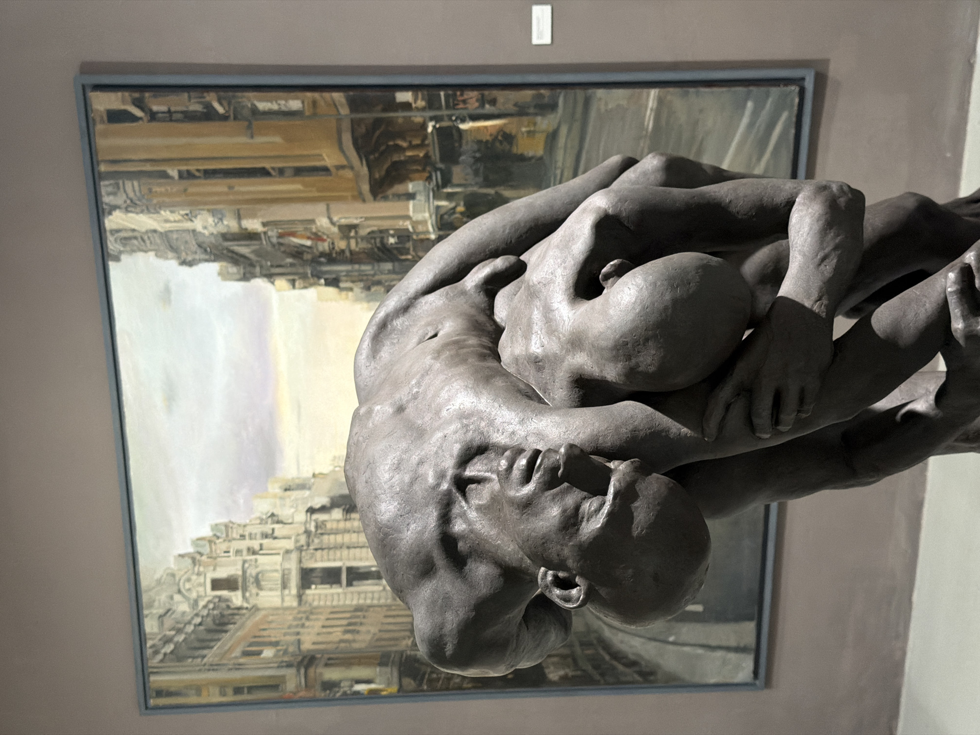 This media file showcases a sculpture of two intertwined figures, rendered in a dark, matte material, positioned in front of a framed painting. The sculpture dominates the foreground, its figures appearing to be in a state of deep embrace or struggle. The figures are highly detailed, with visible musculature and facial features suggesting intense emotion, possibly pain or despair. The composition of the sculpture is dynamic, with limbs and torsos contorting in a complex arrangement.
Behind the sculpture, the painting depicts an urban landscape, likely Barcelona, Spain, as suggested by the context. The painting features a densely packed cityscape with buildings characterized by their intricate architectural details, including balconies and windows. The color palette of the painting is muted, with predominantly warm tones of beige and brown, accented by touches of grey and pale blue, possibly indicating a daytime scene under soft light. The painting is framed with a thin, light blue border.
The overall setting appears to be an indoor exhibition space, such as an art gallery or museum, with plain, neutral-colored walls. A small, rectangular white placard is visible on the wall above the painting, which could be a label or title card for one of the artworks. The lighting in the scene is soft and diffused, highlighting the textures of both the sculpture and the painting. There is no visible text directly on the sculpture, and the text on the placard is too small and indistinct to be legible. The image is oriented vertically, with the sculpture appearing upside down relative to the viewer.