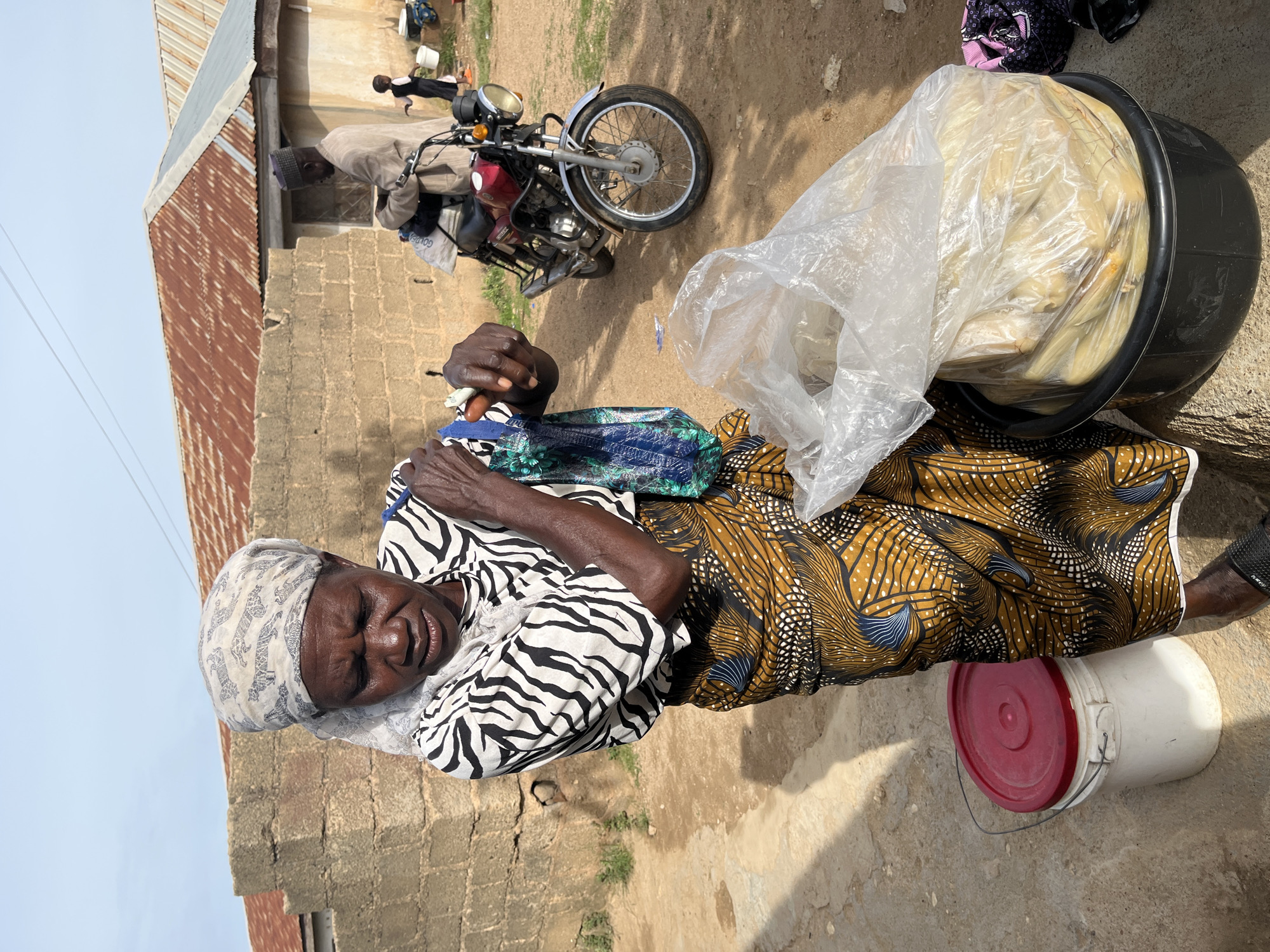 In Rafin Par, Nigeria, a woman is pictured standing outdoors, seemingly engaged in commerce. She is wearing a headscarf and a black and white patterned shirt, along with a vibrantly colored skirt. In her hands, she holds fabric and what appears to be currency, possibly conducting a transaction. In front of her, a large black basin overflows with food items wrapped in plastic, suggesting a food stall or market setting. A white bucket with a red lid sits to her left. In the background, a man in a light-colored robe and cap is seen near a red motorcycle, and a child stands to his right. The environment features rough, sandy ground and buildings with corrugated metal roofs, consistent with rural or semi-urban areas. The overall scene captures a moment of daily life and economic activity.
