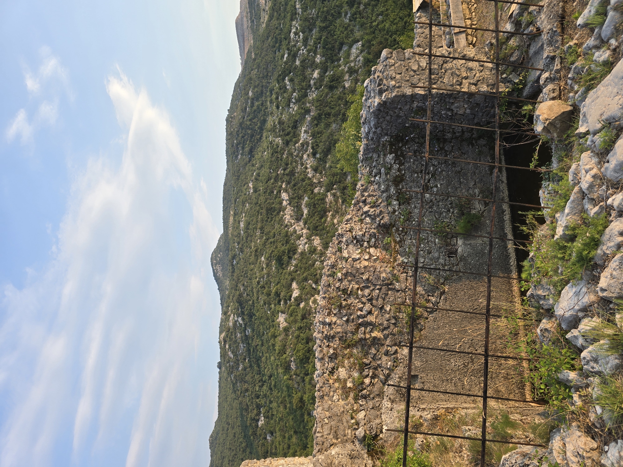 The image captures a view of overgrown hills in Gornja Vrbica, Montenegro, under a partly cloudy sky. In the foreground, there are ruins of a stone structure, possibly an old fortification or building, with a rusty metal fence in front of it. The fence encloses a dark opening, which could be a well or a cistern, partially filled with water or debris. The stonework of the ruins is weathered and shows signs of age, with some vegetation growing on and around it. The surrounding landscape is characterized by rocky terrain and dense green foliage covering the undulating hills that recede into the distance. The lighting suggests it is late afternoon or early morning, with warm tones illuminating the scene. There are no people visible in the image.