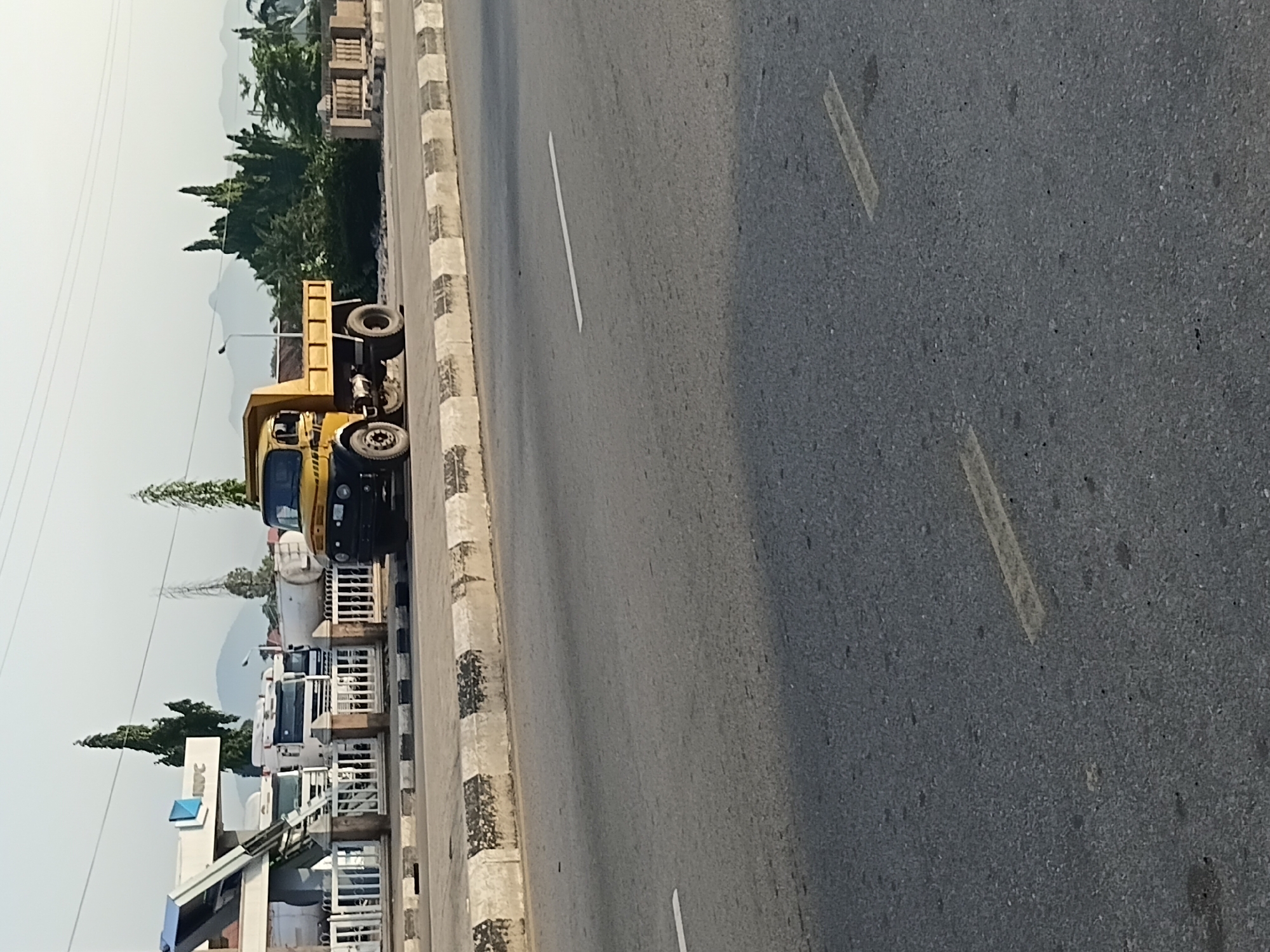 The image captures a street scene in Jalingo, Nigeria, with a prominent yellow dump truck parked on the right side of a paved road. The truck is positioned near a curb with alternating black and white painted markings. In the background, on the left, is a white building with a blue and white logo, appearing to be a fuel station or industrial facility. Several large white trucks, possibly fuel tankers, are parked behind a fence in front of this building. Tall, slender trees, characteristic of the region, are visible behind the trucks. The landscape in the far distance features rolling hills under a clear sky. The road itself is asphalt, marked with white lines indicating lanes. The overall impression is of a typical roadside scene in a Nigerian city, likely during daylight hours given the lighting.