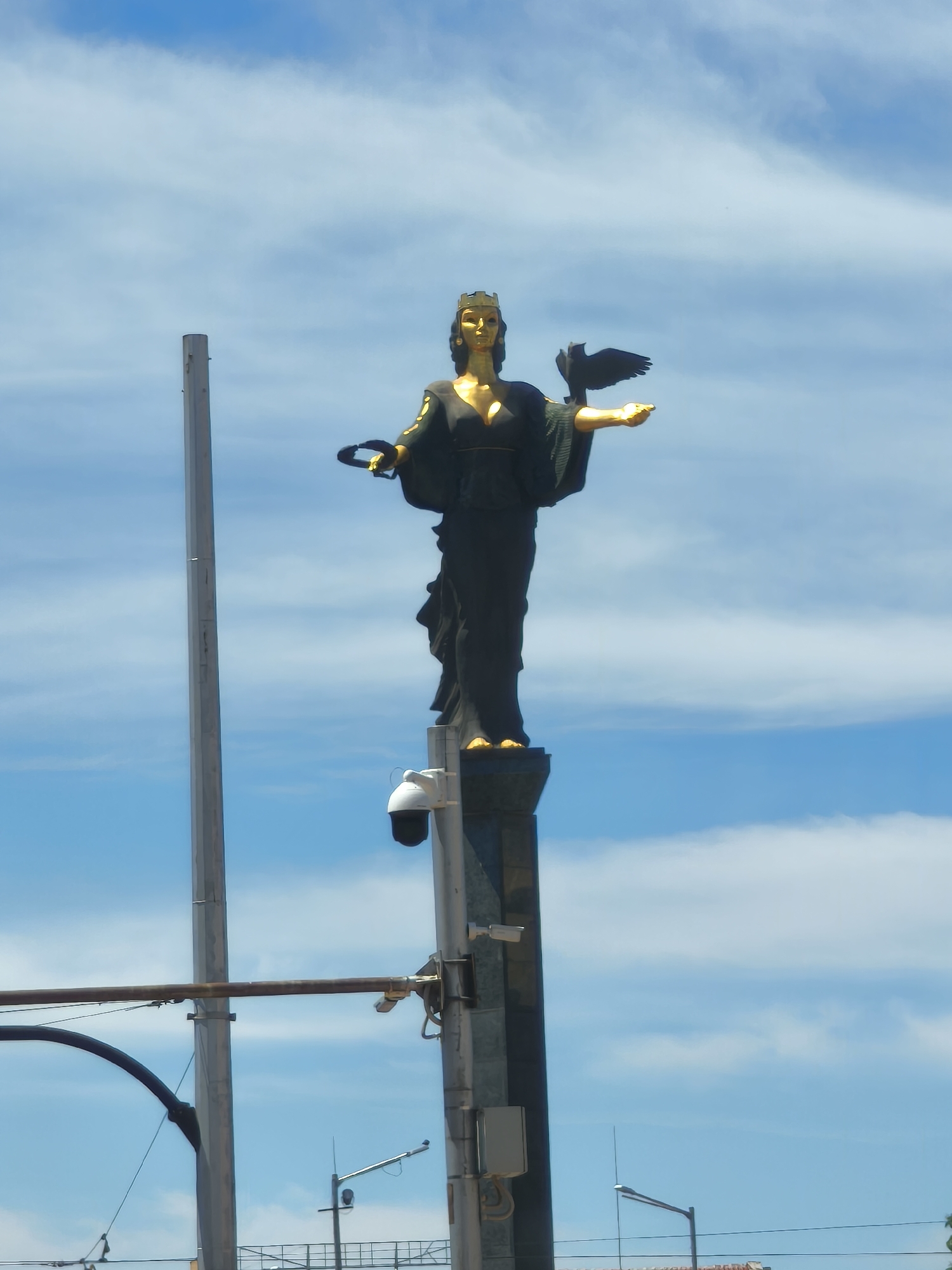 The image shows the Statue of Saint Sofia, a monument in Sofia, Bulgaria. The golden statue depicts a woman holding a wreath in her left hand and a bird on her right. She is standing on a pedestal that also has surveillance cameras attached. Behind the statue is a blue sky with scattered clouds. Power lines and street lights are visible in the background, along with vertical poles and equipment attached to them.