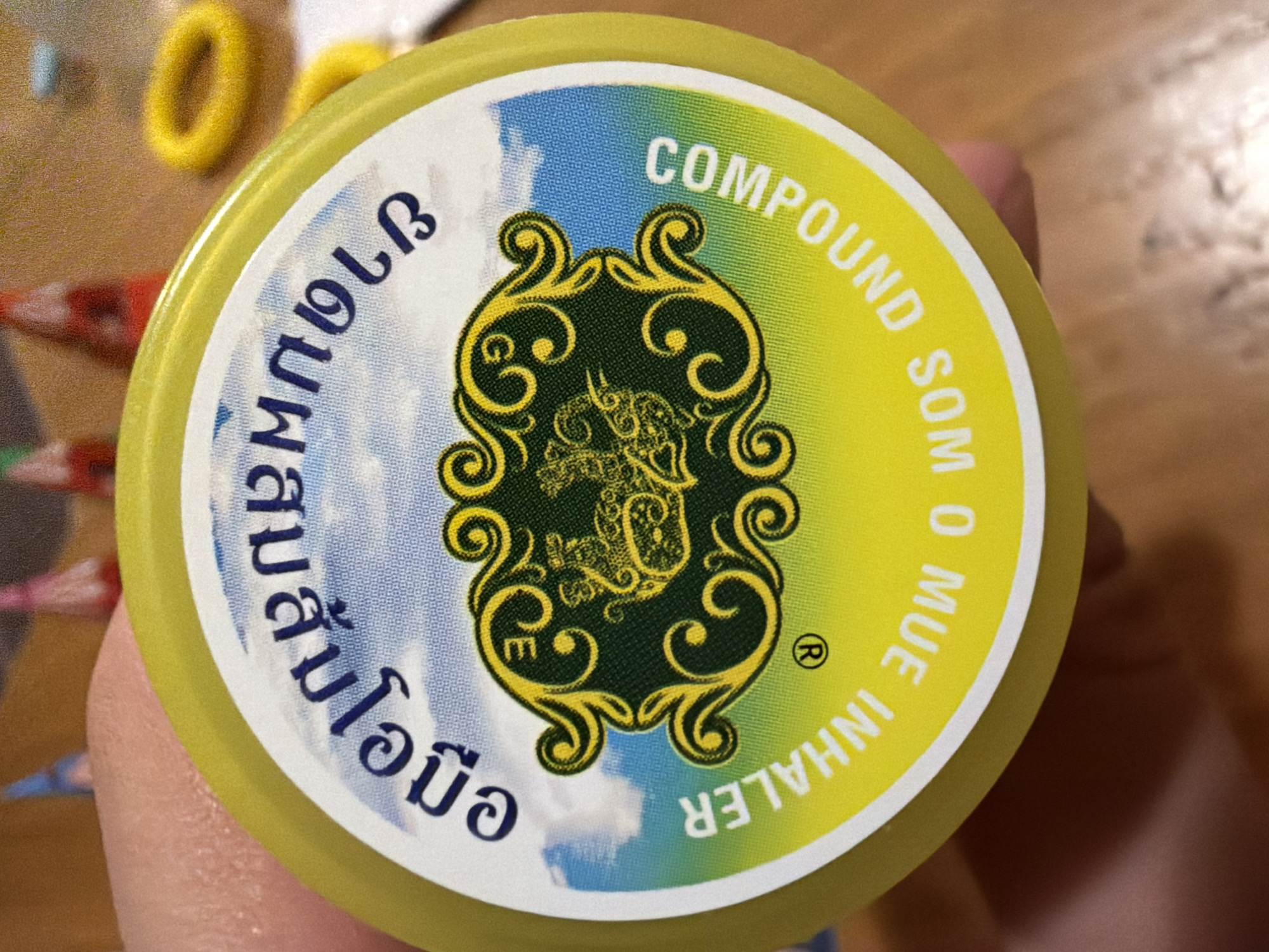 An eye-level close-up shows a yellow jar with a white label, containing the words "COMPOUND SOM O MUE INHALER" in English. The opposite side of the label is inscribed in Thai characters. The label is divided into a blue and white sky pattern and a yellow section. A green ornate design with a stylized animal crest is in the center. The jar is being held in a person's hand. There is no evidence in the image to indicate this is located in Budva, Montenegro.
