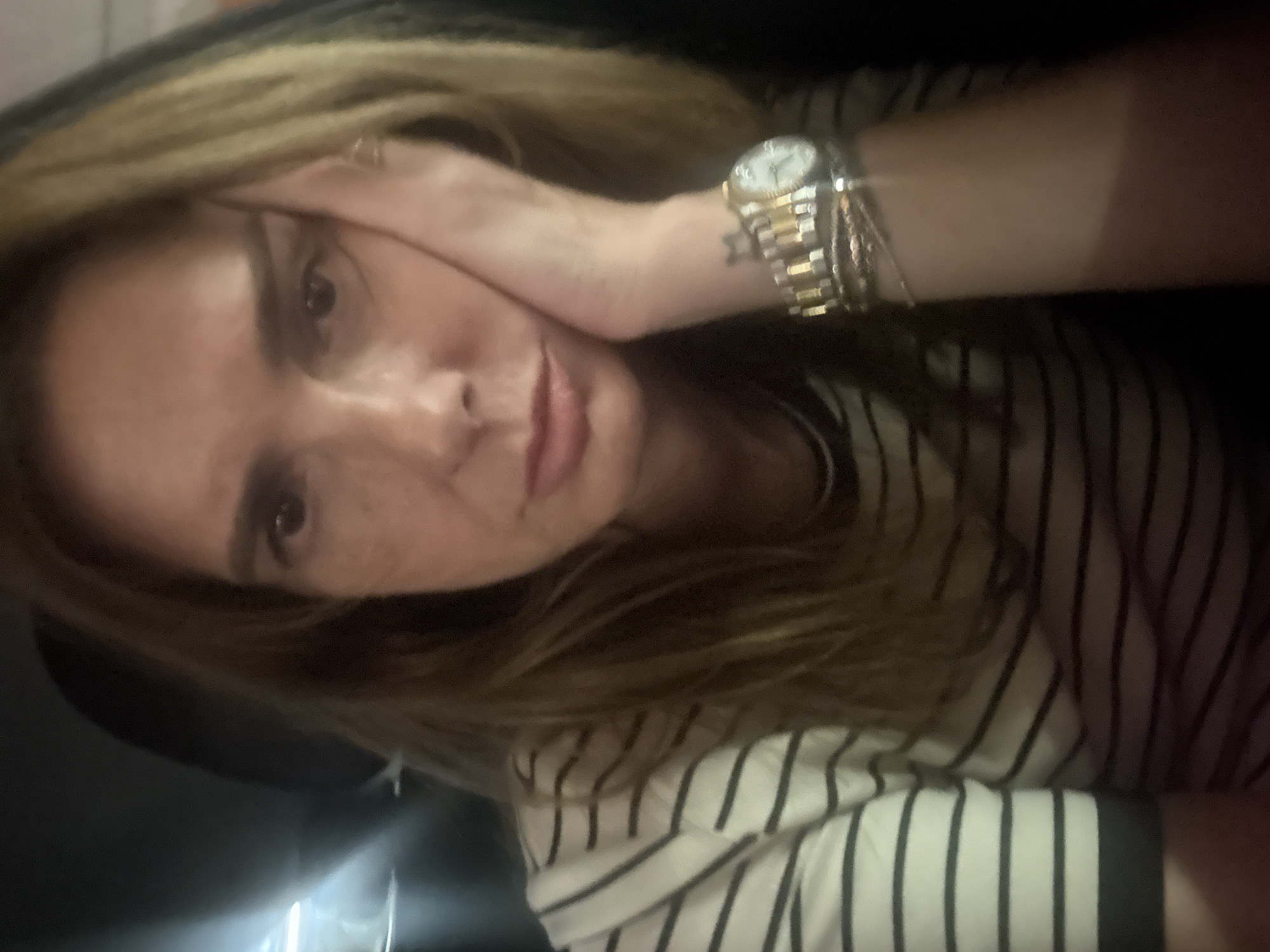 This close-up, rotated portrait shows a woman in what appears to be a dimly lit indoor setting, potentially in Loures, Portugal. She has light brown or blonde hair and is wearing a white top with narrow black horizontal stripes. Her right hand is raised, with fingers resting on her forehead or temple, partially obscuring her face.
On her right wrist, she wears a two-tone (gold and silver) wristwatch with a white face, along with several thin bracelets. Her right eye, appearing dark, is partially visible, as are her slightly parted lips. Faint freckles are visible across her nose and cheeks. The background is dark and out of focus, offering no specific location cues beyond the implied indoor environment.
The activity appears to be a casual moment, possibly a selfie, with the woman striking a relaxed or slightly pensive pose. No specific time of day is discernible, but the lighting suggests artificial light or evening/night. While a watch is present and a faint tattoo is visible below it on her arm, no text or details on either are clearly legible.