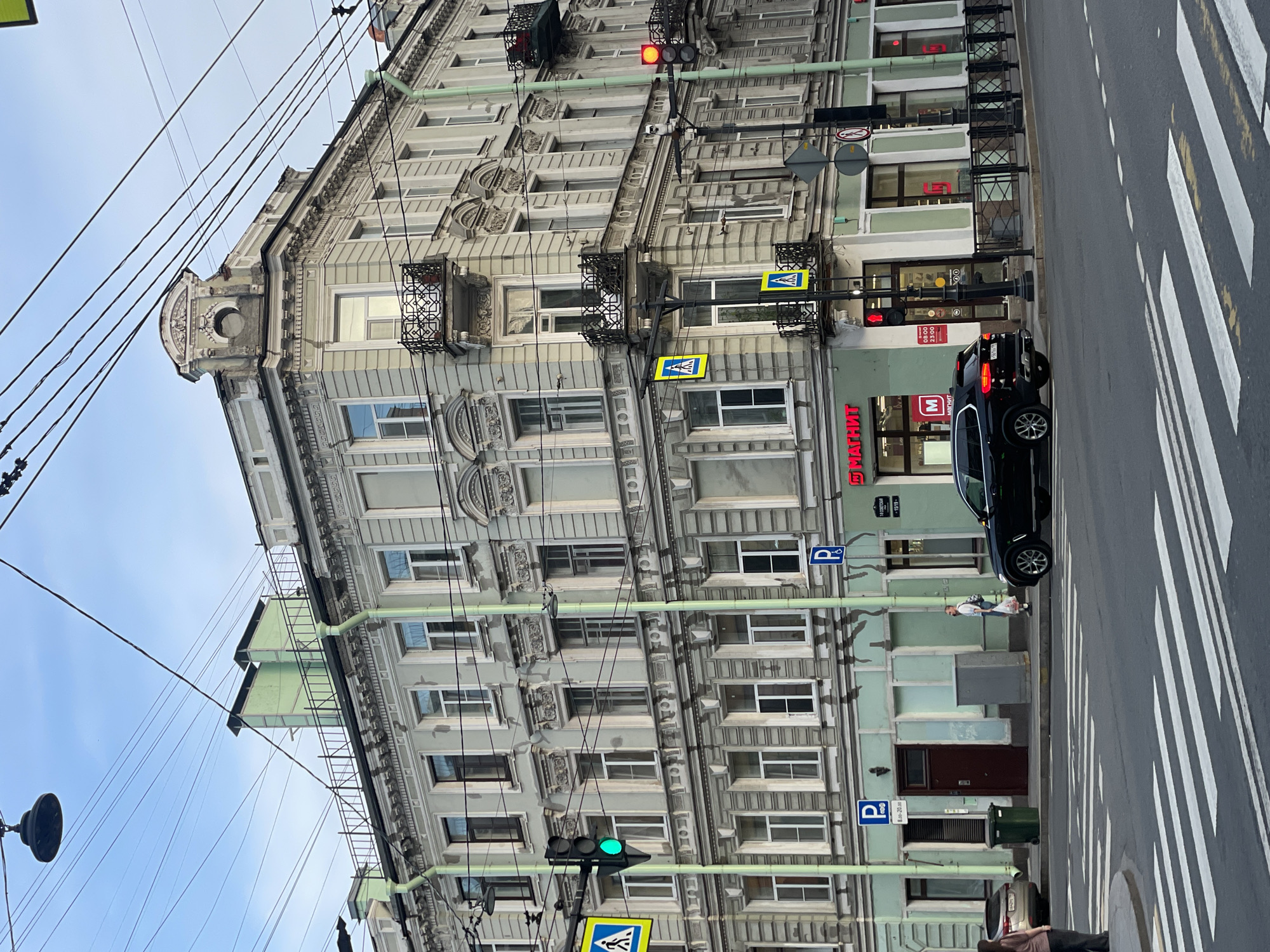 This detailed description is of the intersection of 10-ya Sovetskaya Street and a cross street in Saint Petersburg, Russia.
The image captures a five-story corner building, exhibiting classical architectural elements such as decorative cornices, window surrounds, and balustraded balconies. The facade is predominantly light gray, with the ground floor featuring a mint green lower section. Mint green drainpipes extend vertically down the building. At the building's corner roofline, an ornate, pediment-like structure with a circular recess is visible. On the rooftop, two prominent, light green, angular structures, possibly ventilation shafts, rise above a black railing. Numerous black utility wires crisscross the pale blue, partially overcast sky above the building.
The ground floor houses commercial establishments. The corner unit and the section to its right are occupied by a "МАГНИТ" (Magnit) supermarket, identified by its red signage. The store entrance is visible with a red traffic light above it. To the left of the corner, another entrance, a dark brown wooden door, is situated next to a green trash receptacle and a gray utility box on the sidewalk.
The foreground features an asphalt street with a clear white zebra pedestrian crossing. A black BMW SUV, with a Russian license plate (indicating the 178 region, Saint Petersburg), is positioned at the intersection, its rear visible. To the far left, a silver or beige sedan is partially visible, parked curbside.
Various street infrastructure elements are present. A traffic light on the left is green, indicating go for vehicles, while another on the right is red. Several blue pedestrian crossing signs (a white walking figure within a blue square) are affixed to poles. Blue "P" parking signs, some indicating specific hours (e.g., "8.00 - 22.00"), are visible on poles and affixed to the building. A black metal fence delineates the sidewalk from the street in front of the Magnit store. Other street signs, including a red-bordered triangle (give way) and a red-circled diagonal line (no stopping/parking), are also present.
Two individuals are visible: one person stands partially obscured on the far left sidewalk, and a male pedestrian wearing a light shirt and dark pants walks rightward on the sidewalk near the green trash bin, carrying a white plastic bag. The lighting is diffused, suggesting an overcast day or the softer light of early morning or late afternoon.