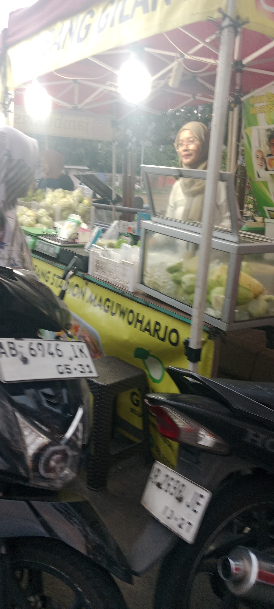 The image depicts a street food stall, likely in Ngemplak, Indonesia, with a focus on fruits and vegetables for sale. A woman wearing a hijab and glasses stands behind the stall. In the foreground, two motorcycles are visible, one on the left with a license plate reading "AB 6946 JK" and another on the right with a license plate reading "JR 2698 JE". The stall itself has a bright yellow awning with text, and displays various produce, including what appears to be guavas and possibly other fruits, in crates and on a scale. The environment suggests an outdoor setting, possibly a marketplace or roadside vendor. The lighting is bright, indicating daytime.