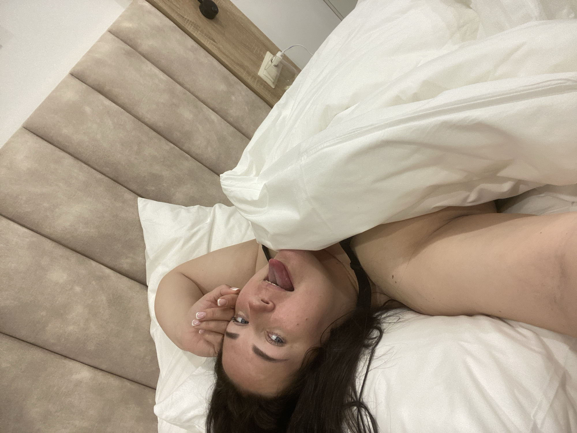 A person lies in bed, with a portion of a white duvet pulled partially over their head. The person, with dark hair and blue eyes, has their tongue sticking out. Their left hand is raised near their face, showing nails with a white tip design. The background includes a padded headboard, a wooden surface with an electrical outlet, and a white wall. The location appears to be an interior room in a building, potentially a hotel or residence, but cannot be definitively identified as being in Sochi, Russia, based on this single image.