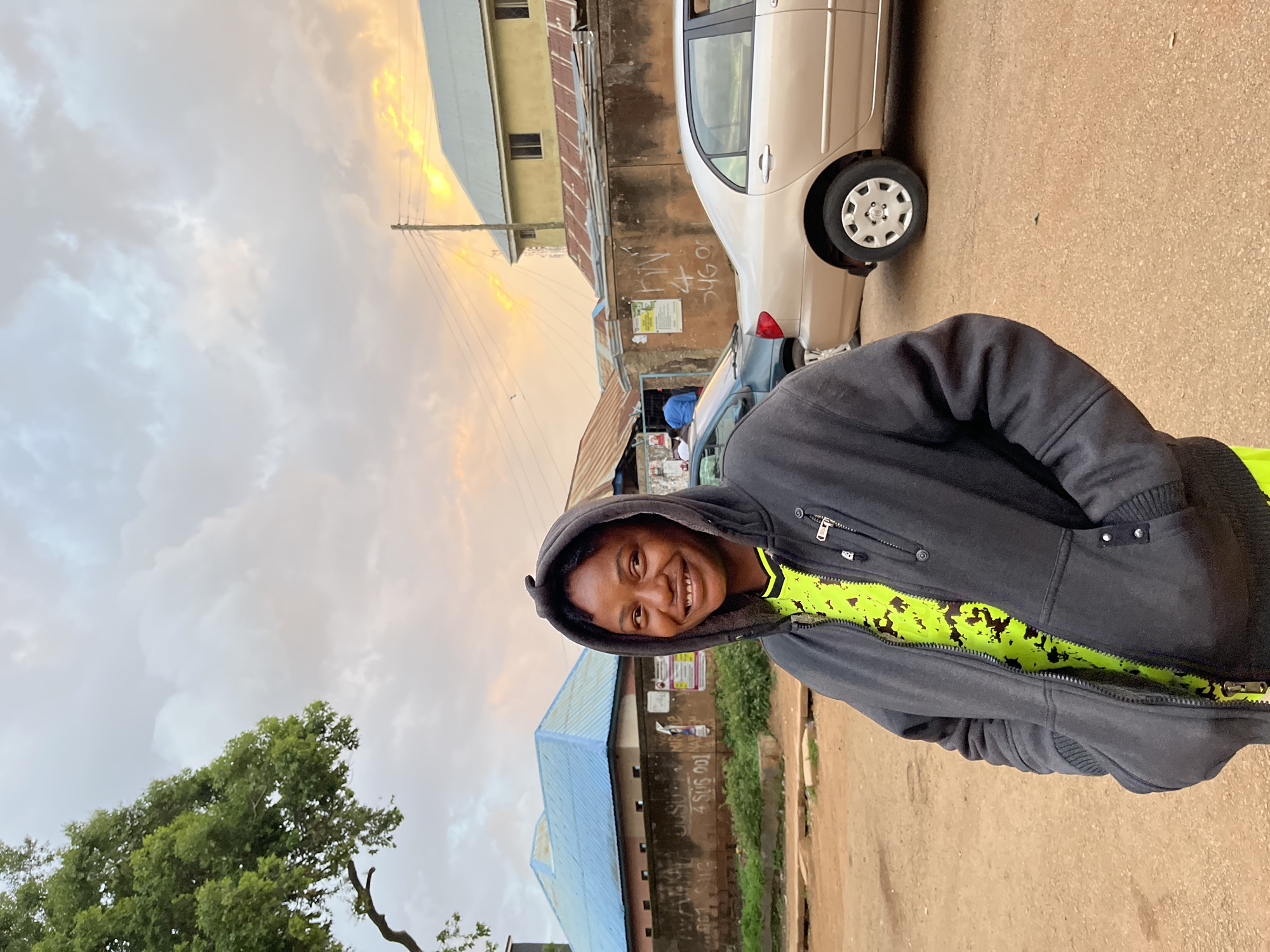 A young Black woman smiles at the camera, standing on a dirt road in Jos, Nigeria. She is wearing a dark gray hooded jacket, zipped down to reveal a bright neon green t-shirt with a black abstract design. Her hands are in the pockets of her jacket. Behind her, a row of buildings with corrugated metal roofs line the street. A light-colored sedan is parked on the right side of the image, and a darker green sedan is partially visible on the left. The sky is overcast with patches of yellow and orange from the setting sun, peeking through the clouds. A large tree with green foliage is visible on the far left of the frame. The overall scene depicts an outdoor urban environment.