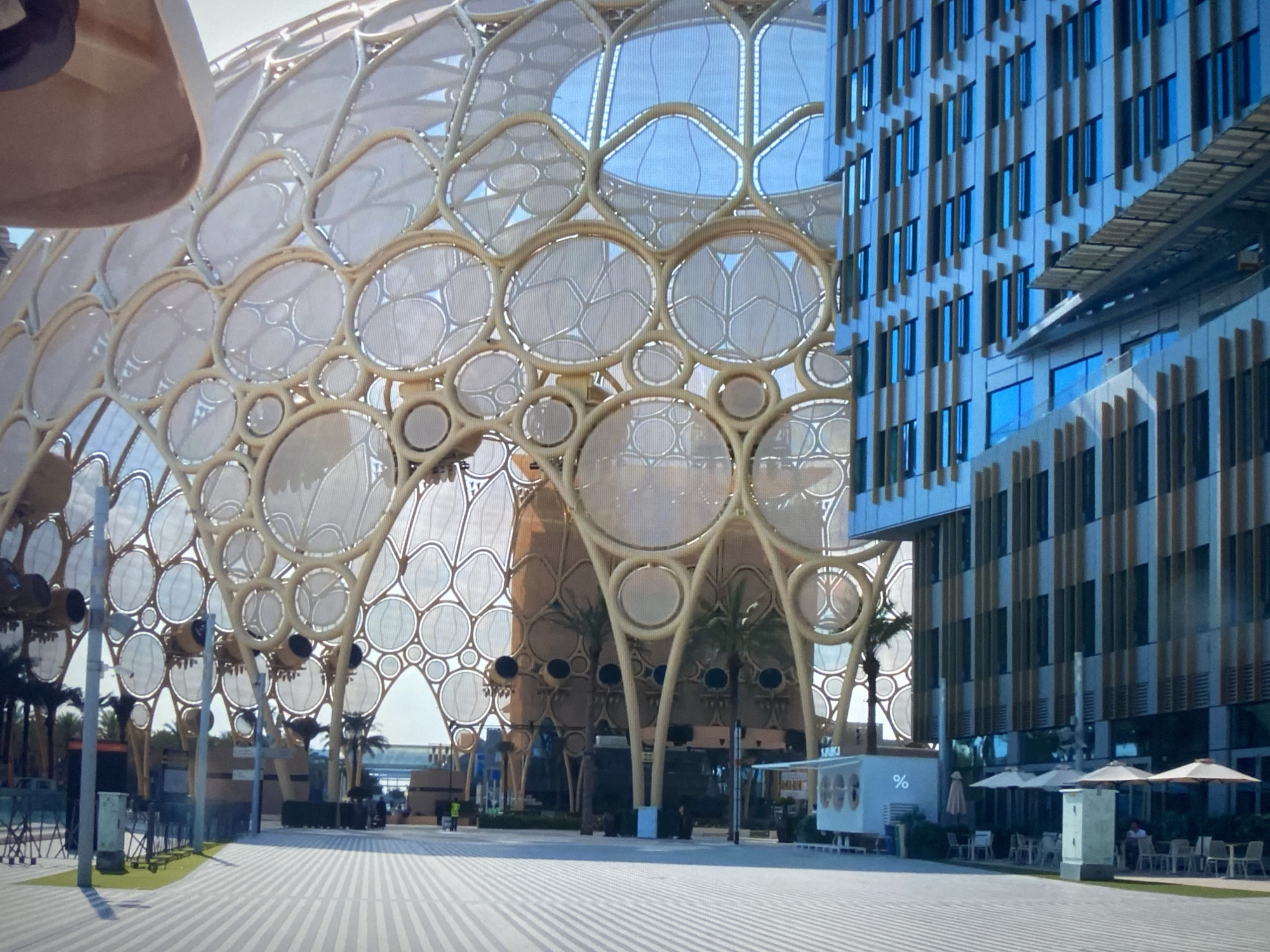 The image shows a modern architectural landscape, likely taken at a public space in an urban setting. On the left, a large, complex structure constructed from a grid of circular and branching elements dominates the scene. It appears to be a pavilion or a large-scale art installation. Palm trees are interspersed amongst the structure, adding a touch of nature. To the right of this structure is a multi-story building with a grid-like facade, featuring many windows and vertical elements. The exterior of the building alternates between reflective glass and solid panels. In the foreground, there's a wide, paved area with linear patterns, suggesting a pedestrian walkway or plaza. A small white booth with a percentage symbol on it is visible on the right, near outdoor seating with tables and umbrellas. The overall scene suggests a contemporary urban design with a blend of architecture, nature, and public space amenities.