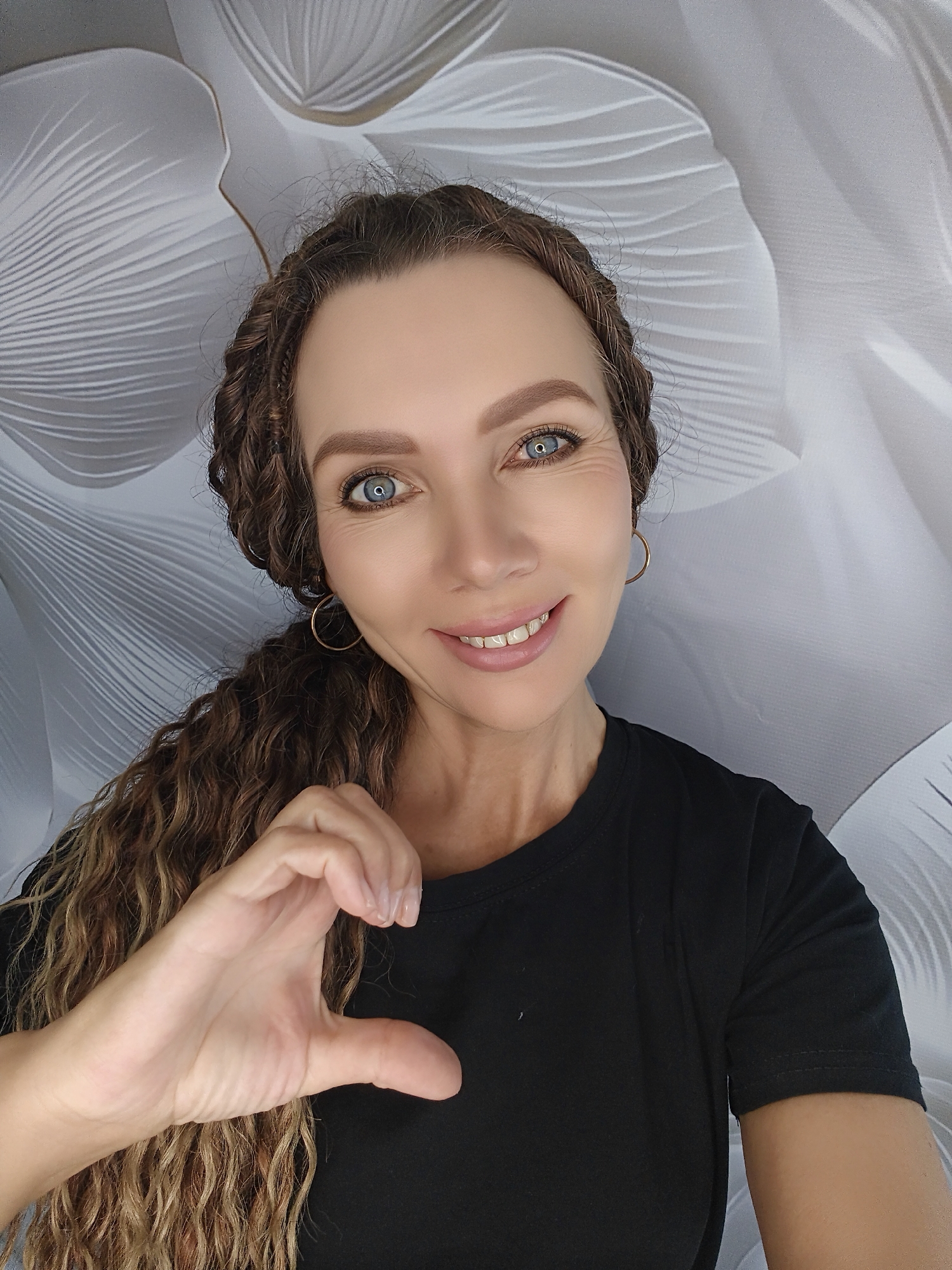 A medium shot shows a woman in a black t-shirt against a white floral background. The woman has long, curly brown hair braided on one side and down the other. She has blue eyes and is smiling with her teeth showing. Her left hand is in the foreground, forming a heart shape with her thumb and index finger. She is wearing gold hoop earrings. The background features large, stylized white floral or petal shapes with visible texture.