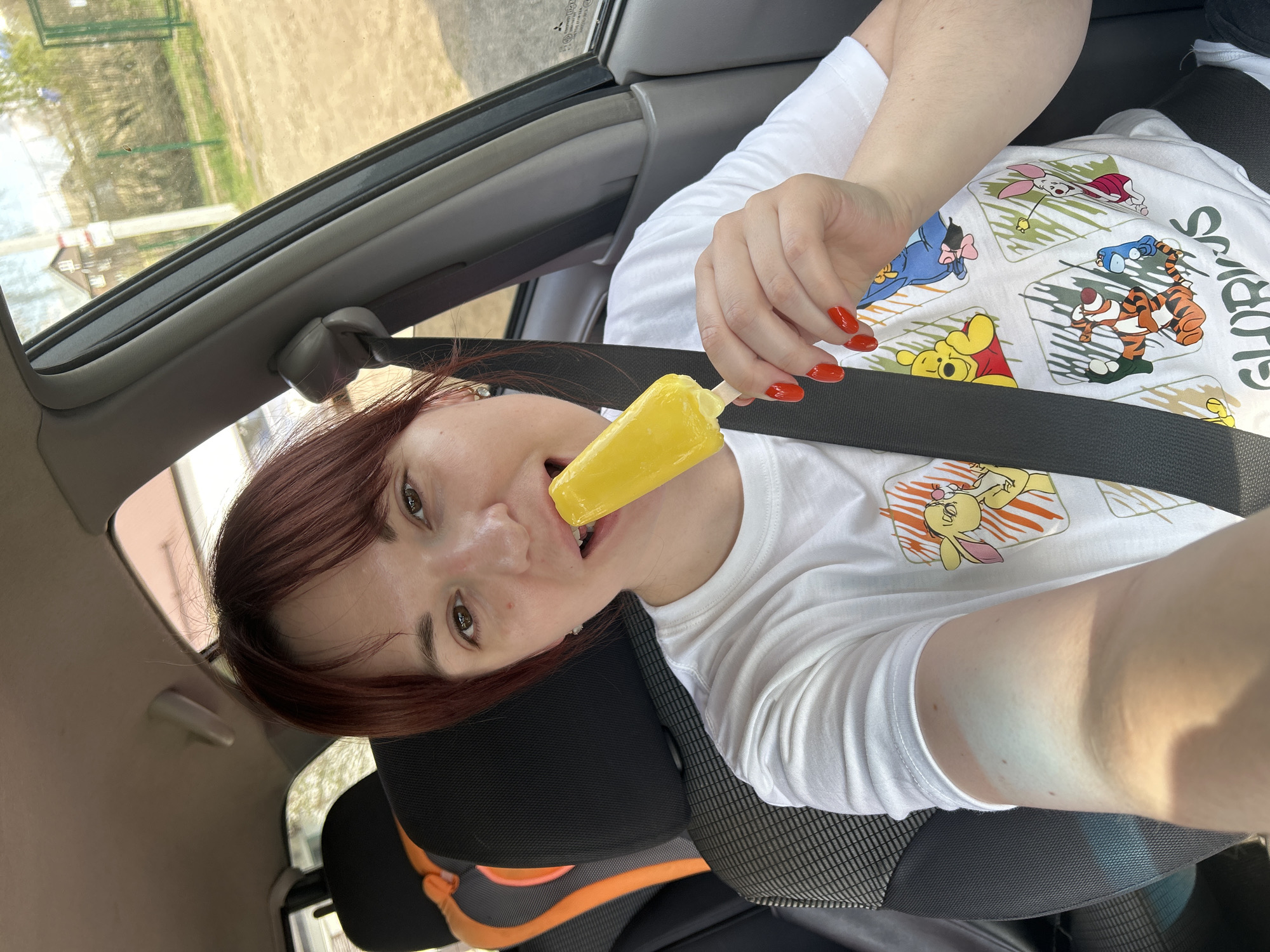 A woman with auburn hair is in a car, wearing a white t-shirt with Winnie the Pooh characters on it, eating a yellow popsicle. She has orange nail polish and is wearing a seatbelt. The background shows a blurred view of the outside. The location, Фоки, Russia is not deducible from this image.