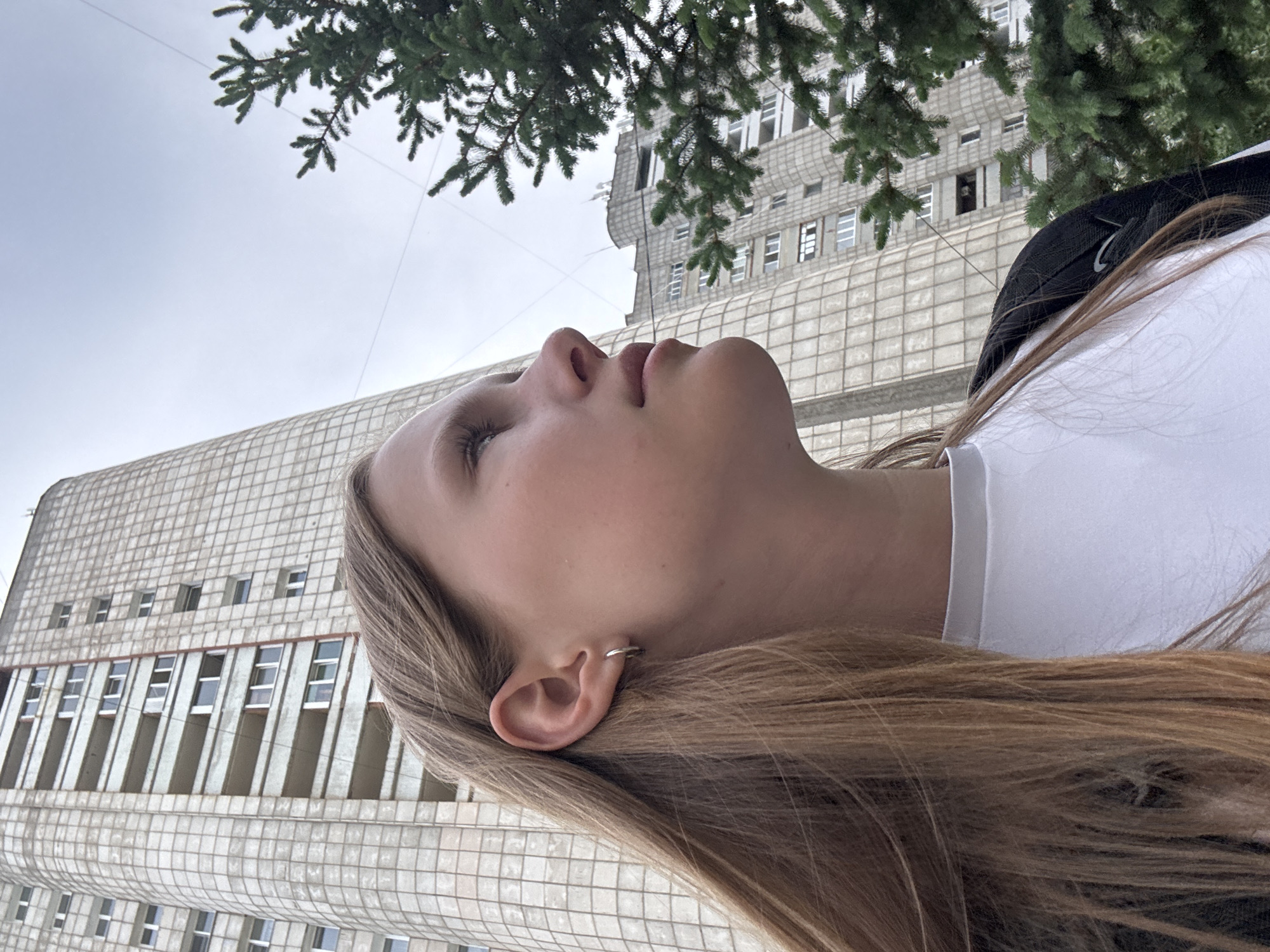 The image shows a young woman in Chaikovsky, Russia. She is wearing a white shirt and a backpack with a black Nike logo. She is facing upwards towards the sky and the top of a building. Her hair is light brown. A tree branch hangs above her. The building is a grid-like structure with rows of windows. The sky is overcast.