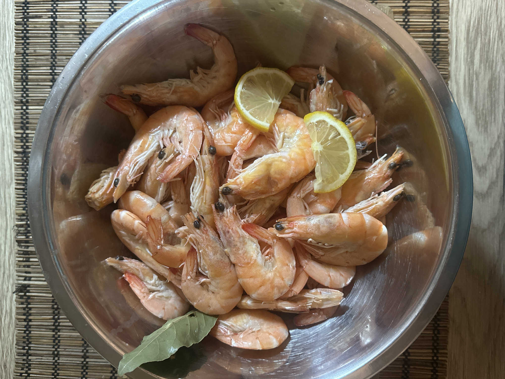 A top-down shot features a stainless steel bowl filled with cooked shrimp, arranged on a wooden placemat. Two lemon wedges are positioned among the shrimp. A single bay leaf rests near the bottom of the bowl. There are no people visible in the image, and the environment appears to be an indoor setting, likely a kitchen. The location in the city of Moscow, Russia cannot be confirmed from the image.