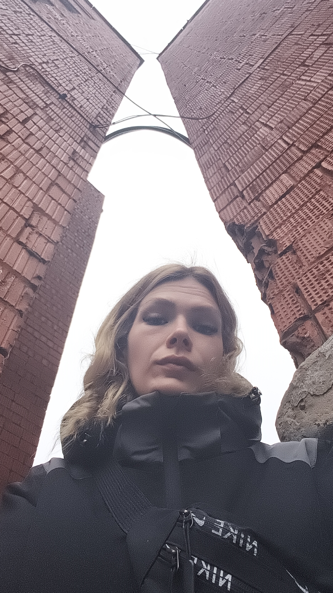 A woman is standing in front of the "Forever with Russia" monument in Izhevsk, Russia. The monument consists of two large brick towers. The sky is white. The woman is wearing a black jacket and a black bag with white lettering that reads "NIKE." The camera angle is from below, looking up at the woman and the monument. The right tower shows signs of significant damage.