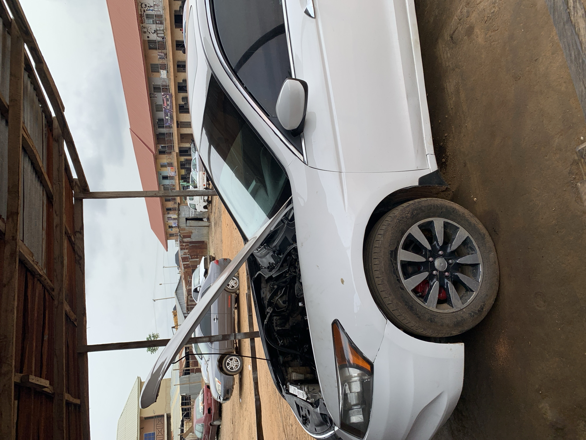An outdoor scene captures a white sedan, likely a Honda Accord, with its hood open, revealing the engine compartment. The car's front right wheel, featuring dark multi-spoke alloy rims and a visible red brake caliper, is prominent in the foreground. The vehicle appears to be in a state of repair or inspection.
The setting is an unpaved, brownish-gray dirt ground, suggesting a workshop or car service area. To the left, a rustic structure constructed from wooden beams and corrugated metal sheets forms part of the immediate foreground, indicating an informal or practical workspace.
In the background, other vehicles, including a gray sedan and parts of red and white cars or vans, are parked. Beyond these, a row of multi-story buildings with varied designs and possibly mixed residential and commercial use can be seen. One building features red-colored upper floors or roof sections and balconies. Some banners or signs are affixed to the buildings, but the text is not legible due to distance and image resolution.
The sky is overcast and gray, suggesting a cloudy day without direct sunlight. The overall environment portrays a functional, busy area, likely a mechanic's or car repair shop, in a semi-urban or developing setting. No individuals are visible in the image. The scene conveys a sense of everyday activity in an automotive service context.